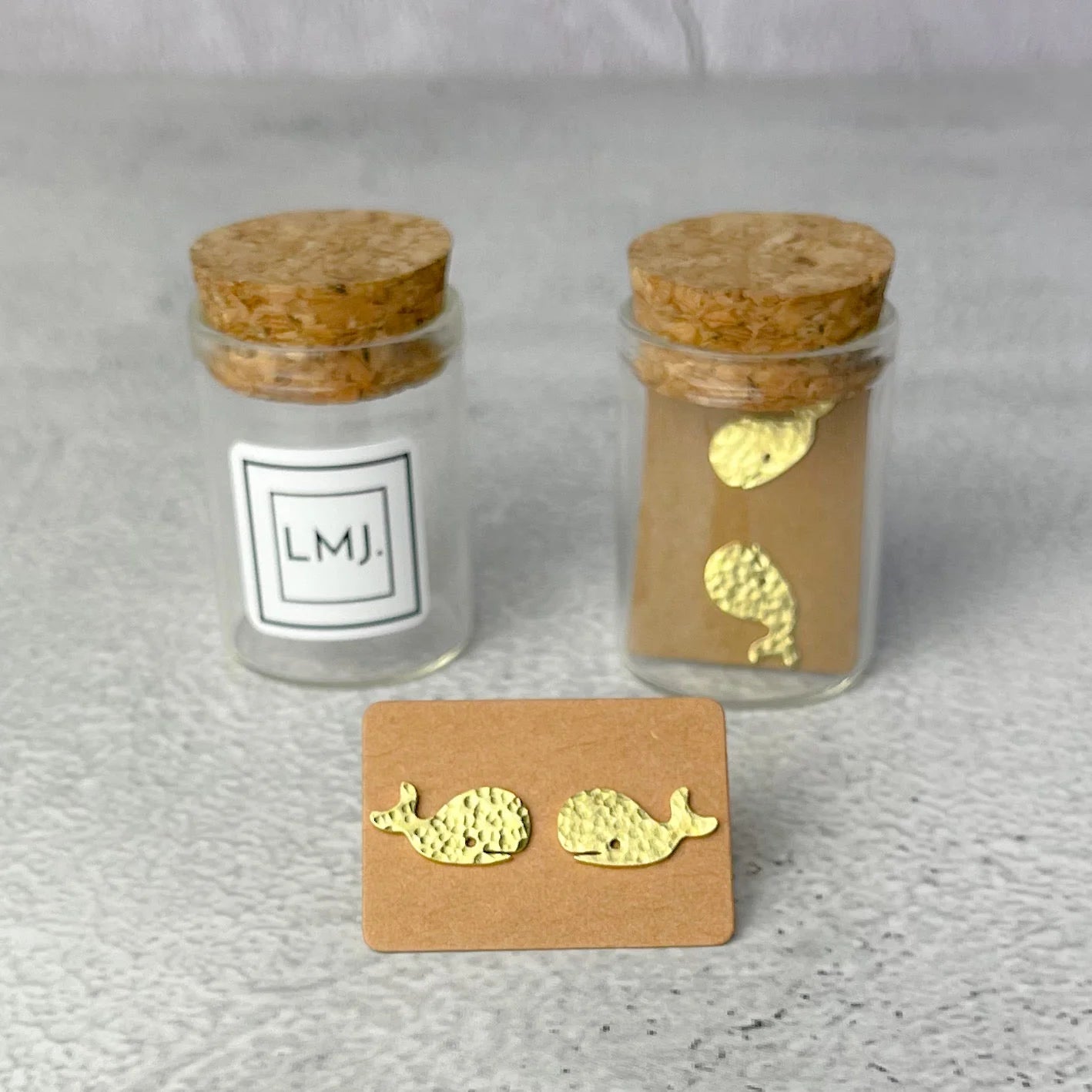 Gold whale earrings on a card with two small glass jars labeled 'LMJ' on a gray background.