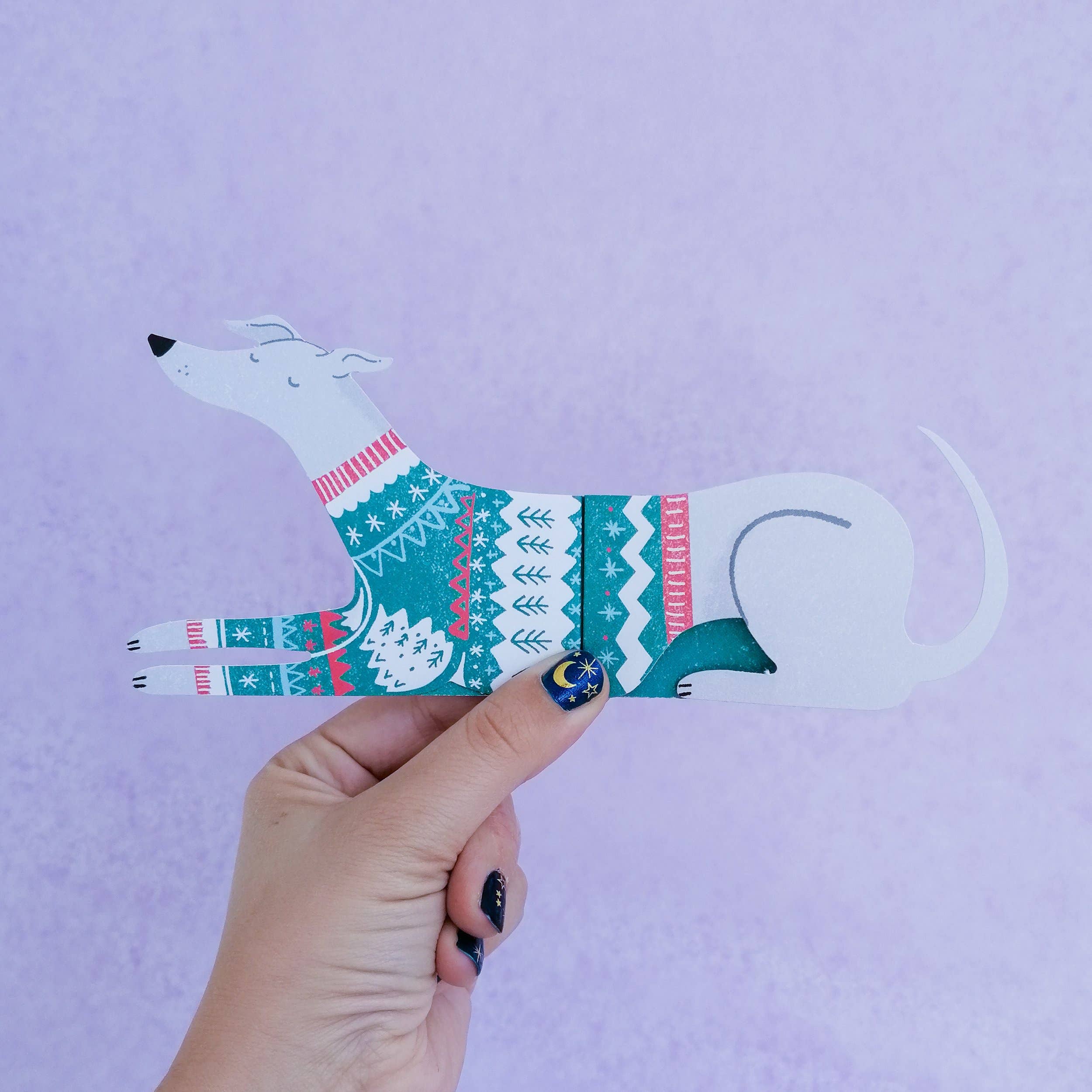 Hand holding a paper cutout of a dog wearing a sweater against a purple background.