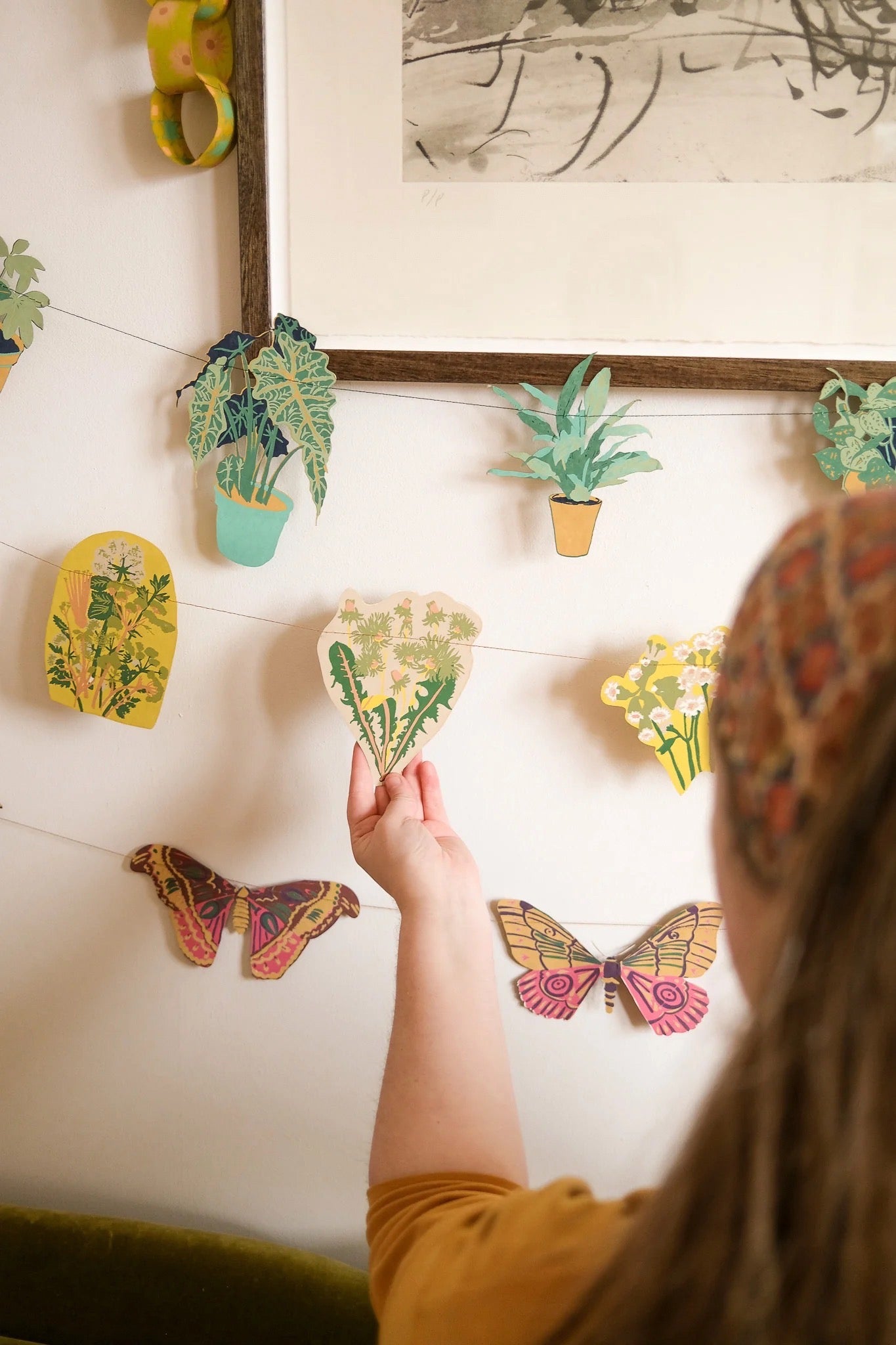 Person arranging decorative paper plant, flower and butterfly garlands on a wall.