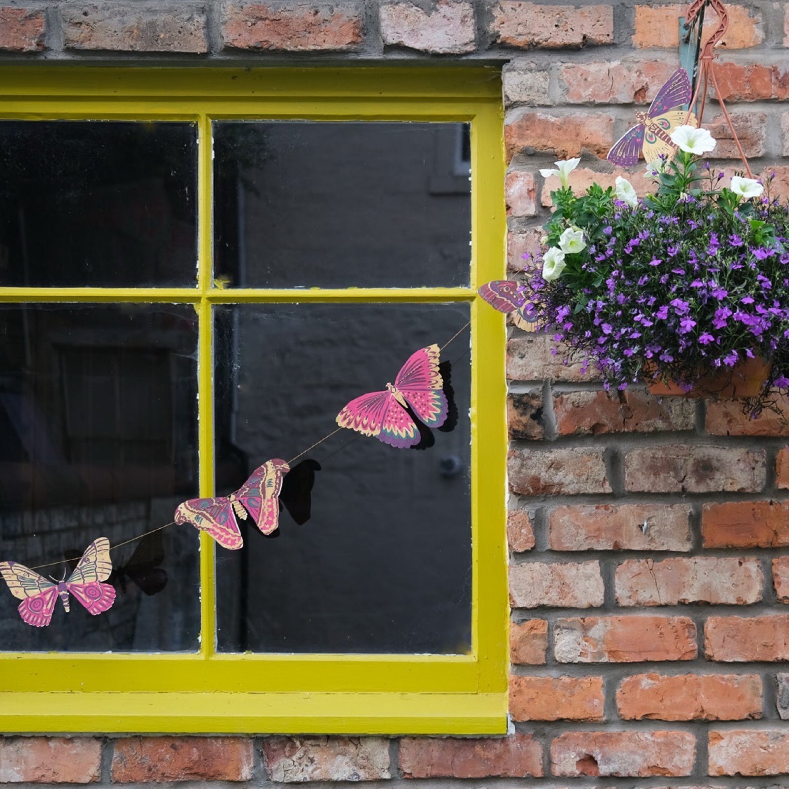 Beautiful butterfly garland, featuring ten screen printed paper butterflies in various pink, red, green and yellow designs hanging against a window with a yellow frame and a brick wall.