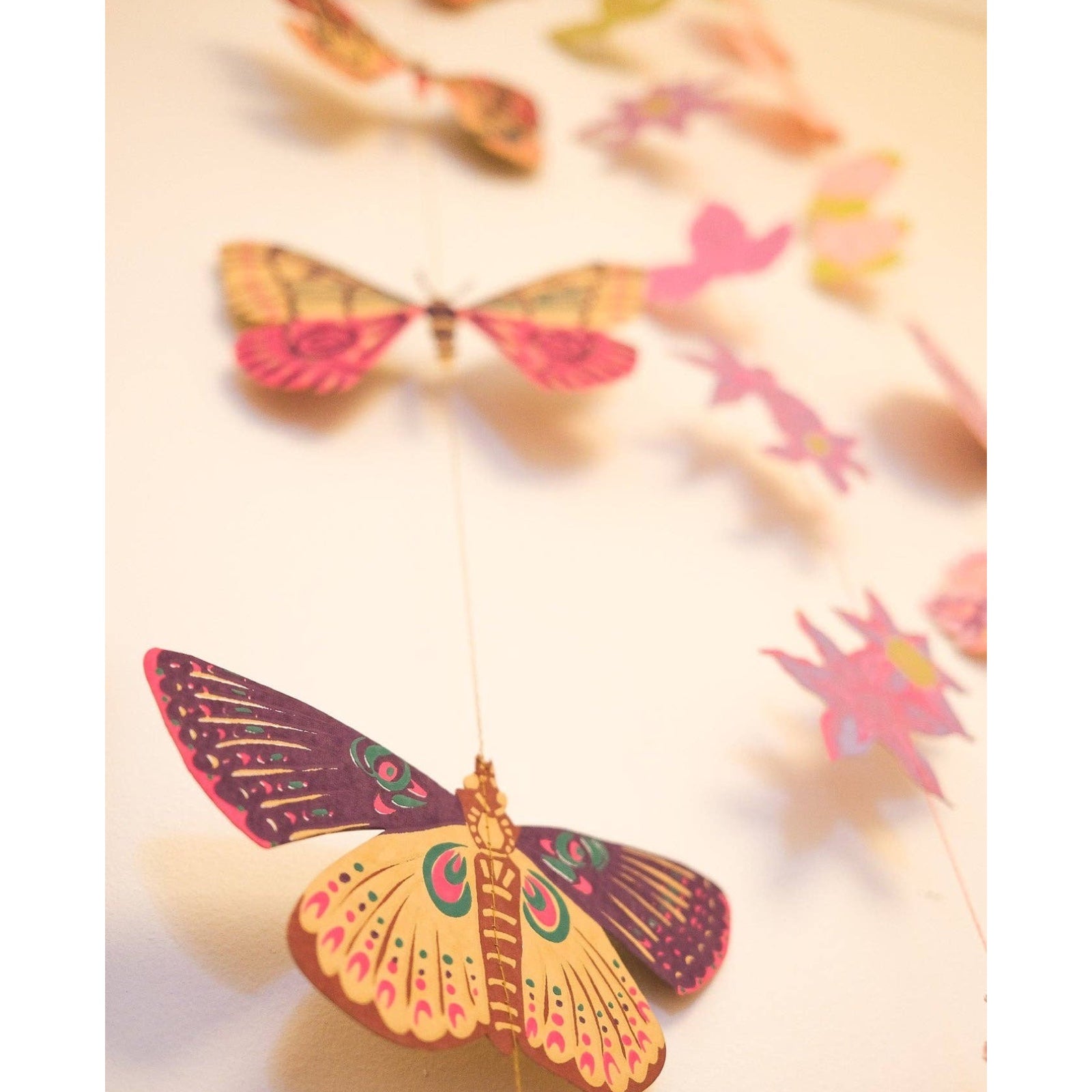 Vertical butterfly garland, featuring ten screen printed paper butterflies in various pink, red, green and yellow designs hanging against a white wall.