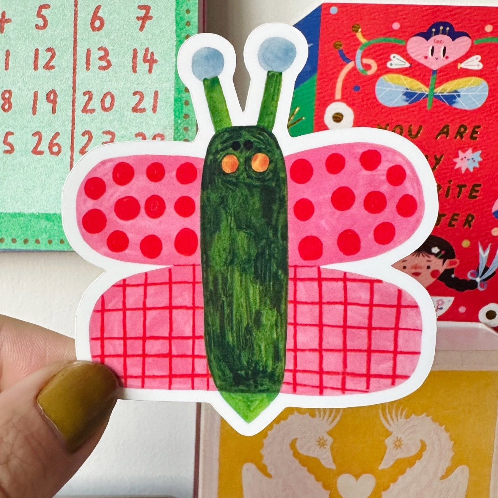 A hand with mustard green nail polish holding a die-cut sticker of a whimsical butterfly. The butterfly has a green body, red polka-dot wings, and red grid-patterned wings, with blue antennae and orange eyes.