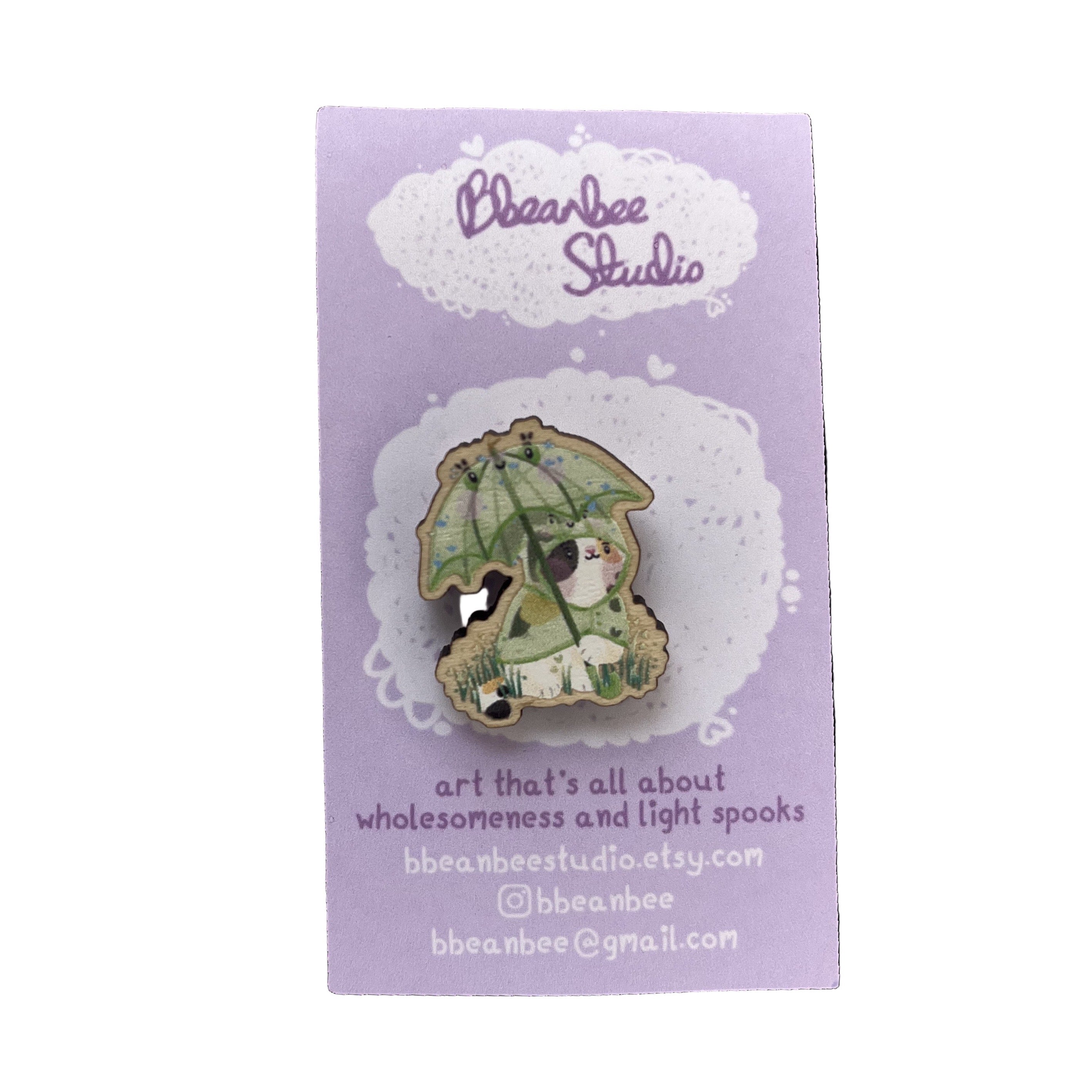 Wooden pin badge of a calico cat wearing a frog raincoat, holding a matching frog umbrella. This badge is printed onto 4mm thick responsibly sourced UK mapelwood. The badge has a pink, heart-shaped, rubber backing.