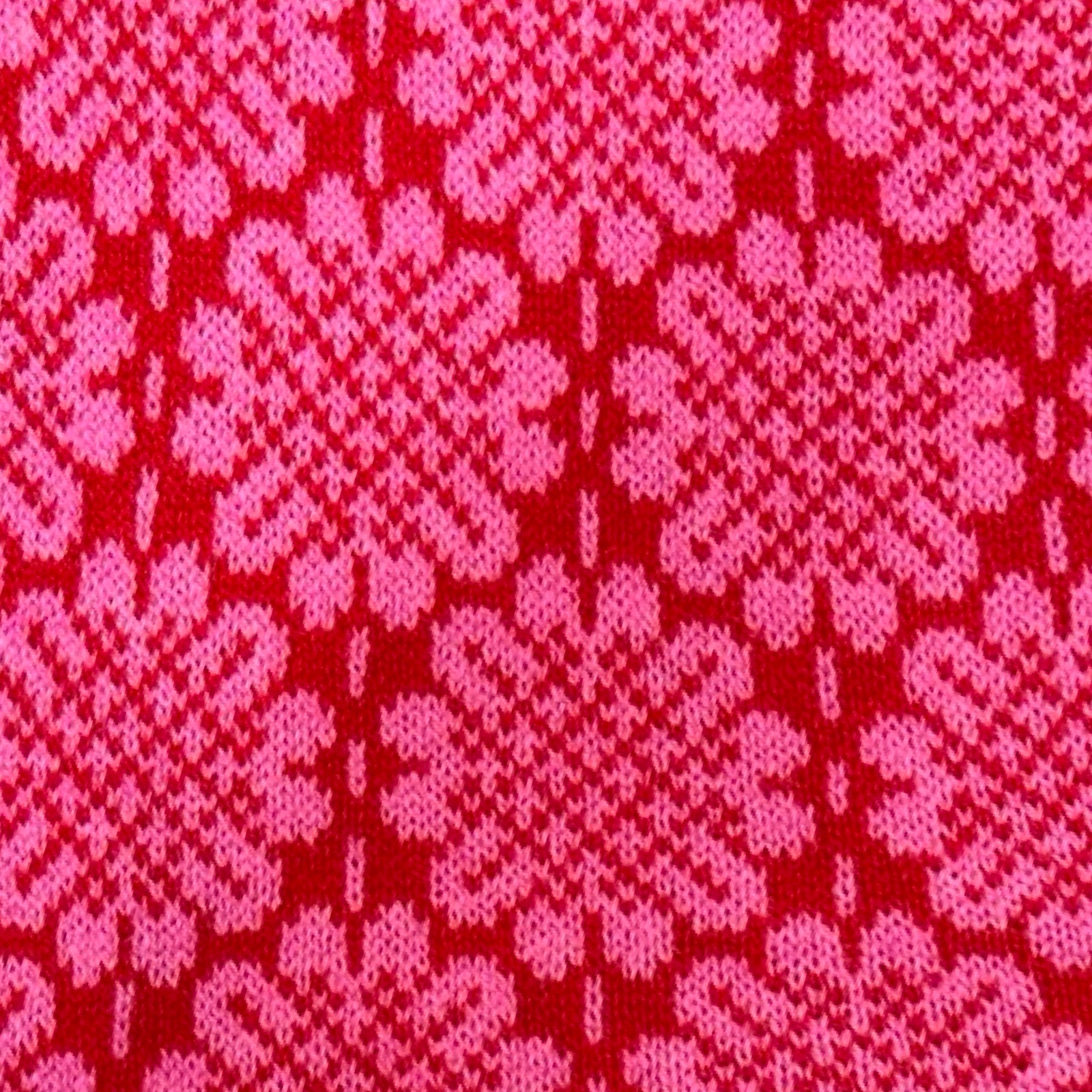 A 100% lambswool knitted scarf with a pink daisy pattern against a red background with contrasting green edges.