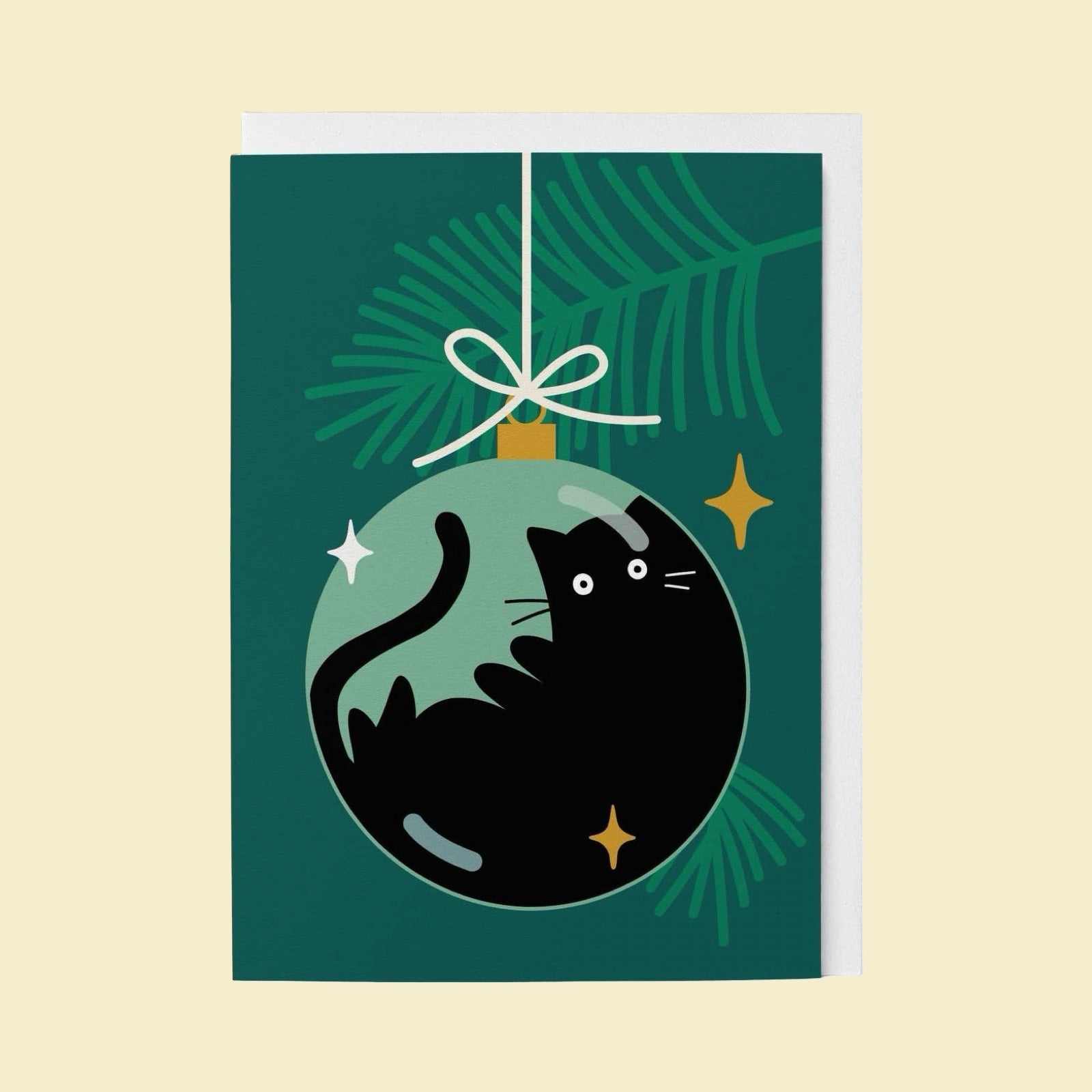 A Christmas greeting card featuring a black cat curled up inside a green ornament, hanging against a dark green pine branch background with gold stars, shown on a light cream backdrop.