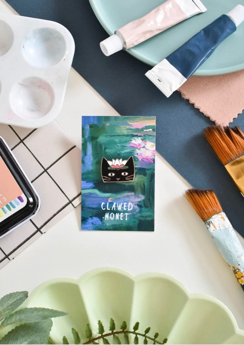 Artistic card with a cat pin surrounded by art supplies on a table.
