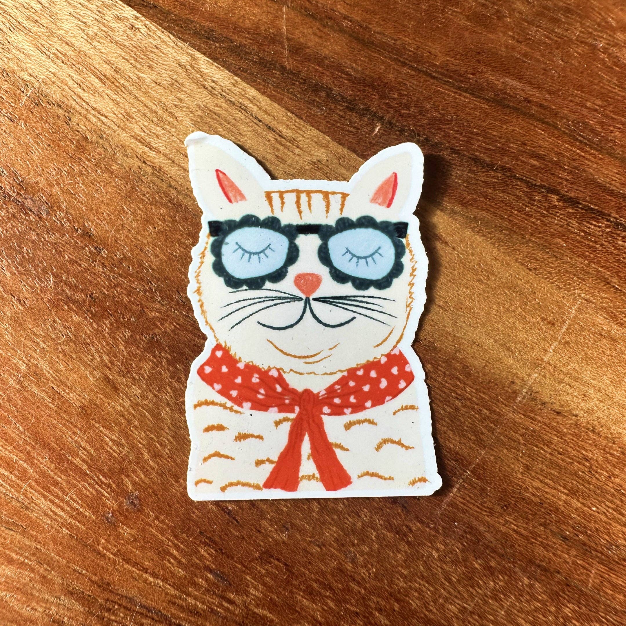 Cat-shaped sticker with sunglasses and a red bow tie on a wooden surface.