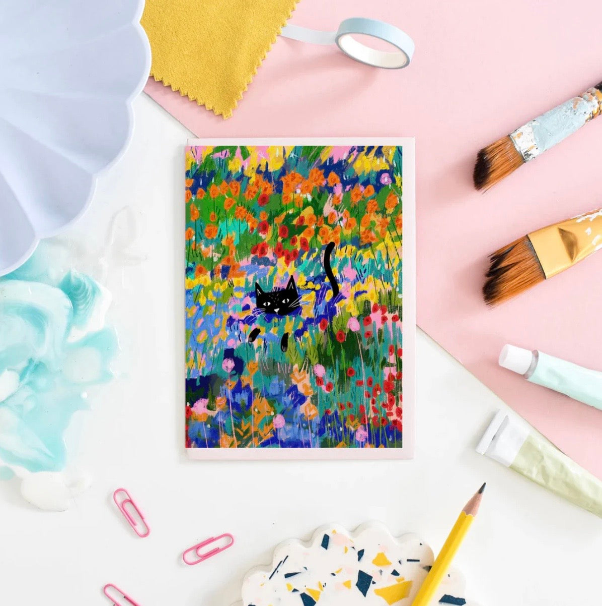 Colourful painting of a cat in a garden with art supplies on a pink background.
