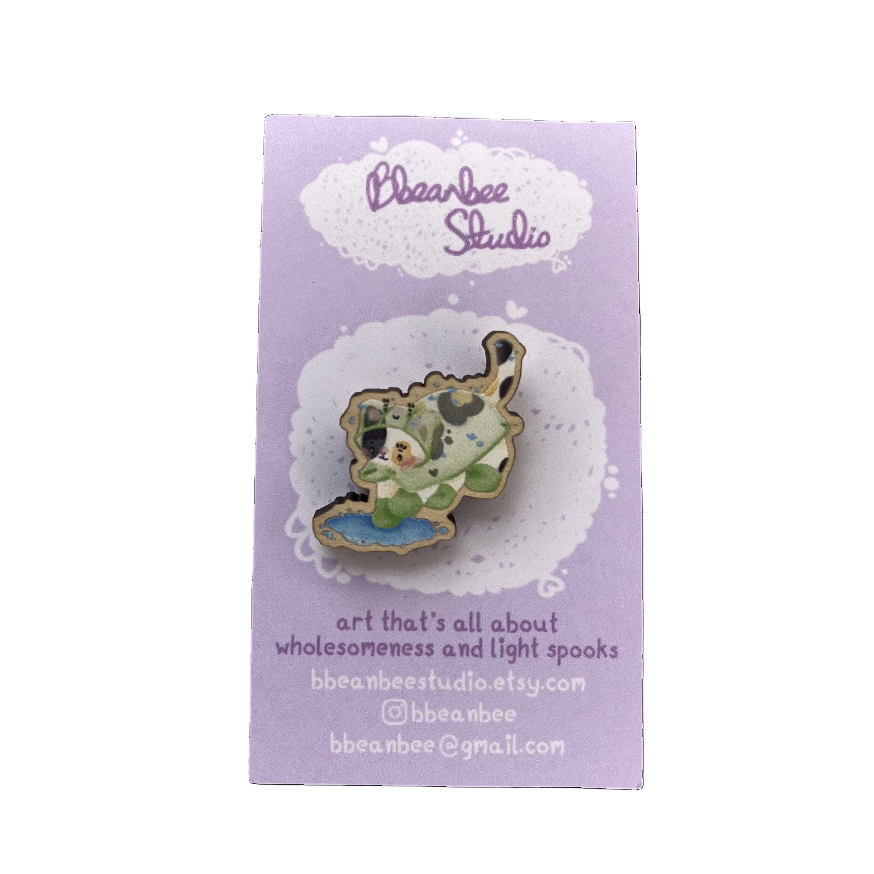 Wooden pin badge of a calico cat wearing a frog raincoat and green rainboots, jumping into a puddle - on a puirple backing card