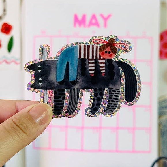Hand holding a decorative cat sticker in front of a colourful calendar and wall decorations.
