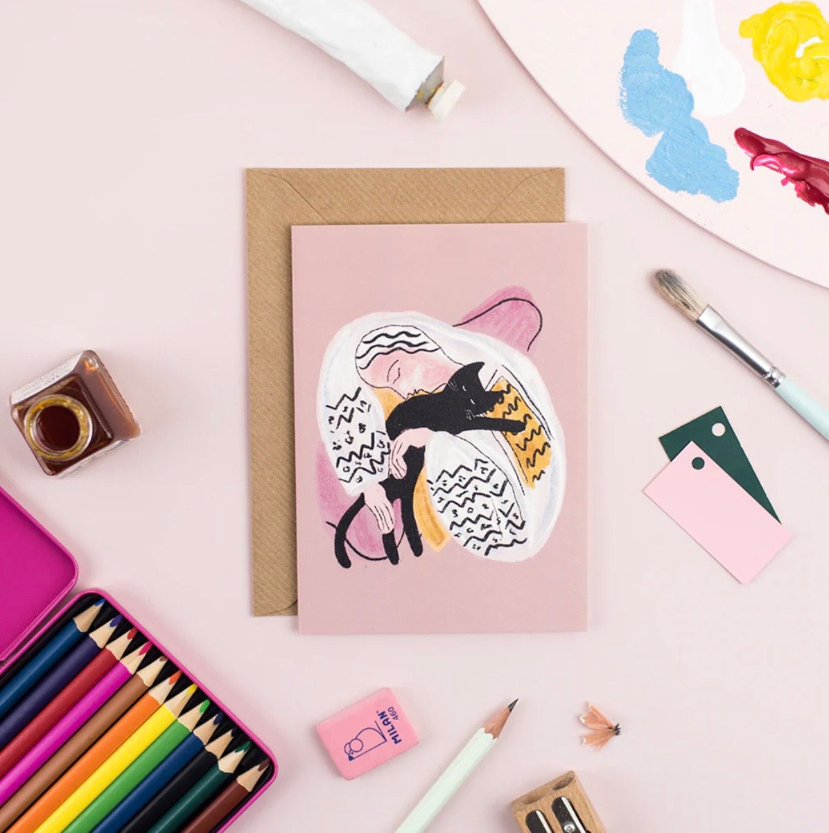 Greeting card with a cat illustration on a pink background, surrounded by stationery items.