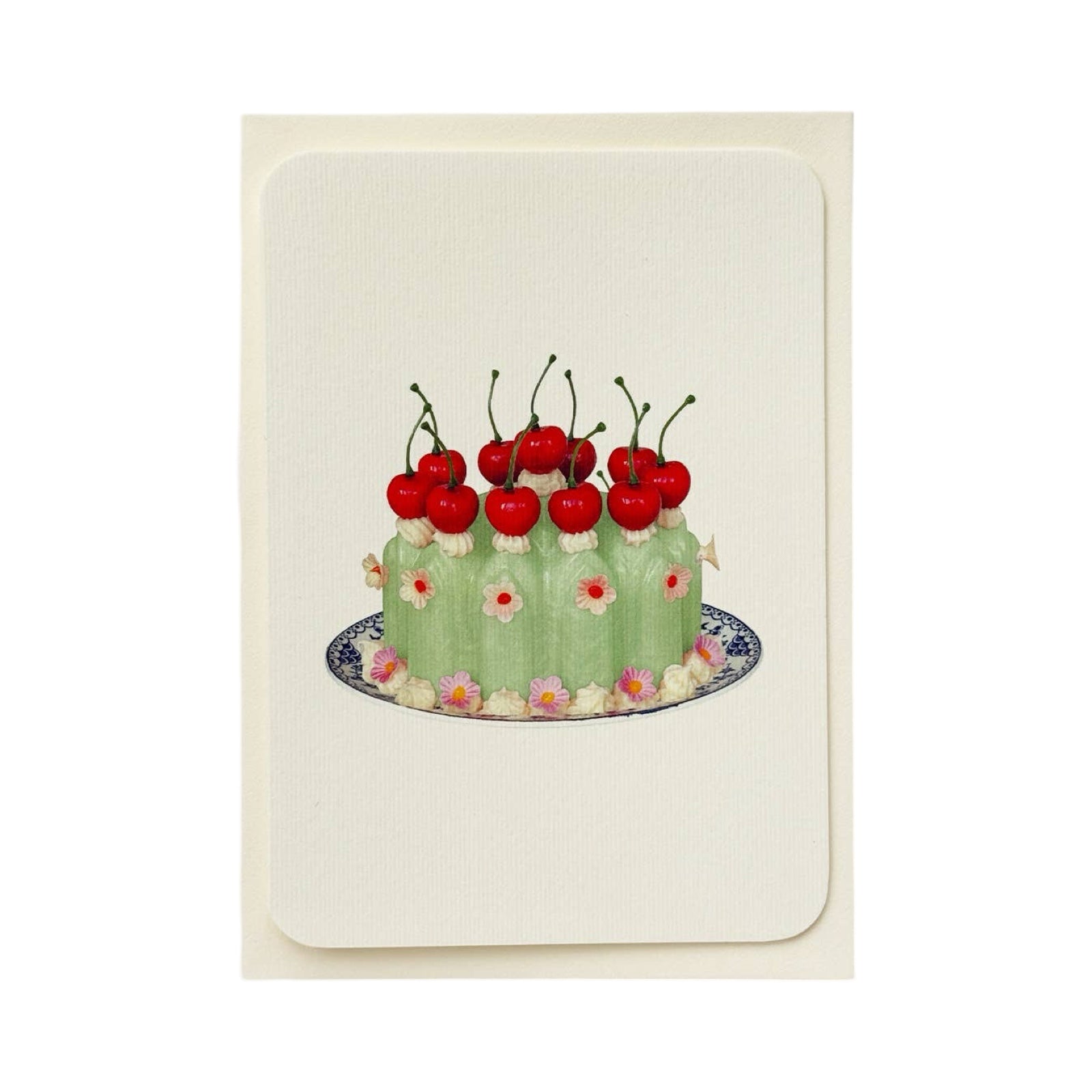 A cream coloured birthday card with a green coloured blancmange with cherries and cream on the top and small pink flowers, all on a blue and white plate.