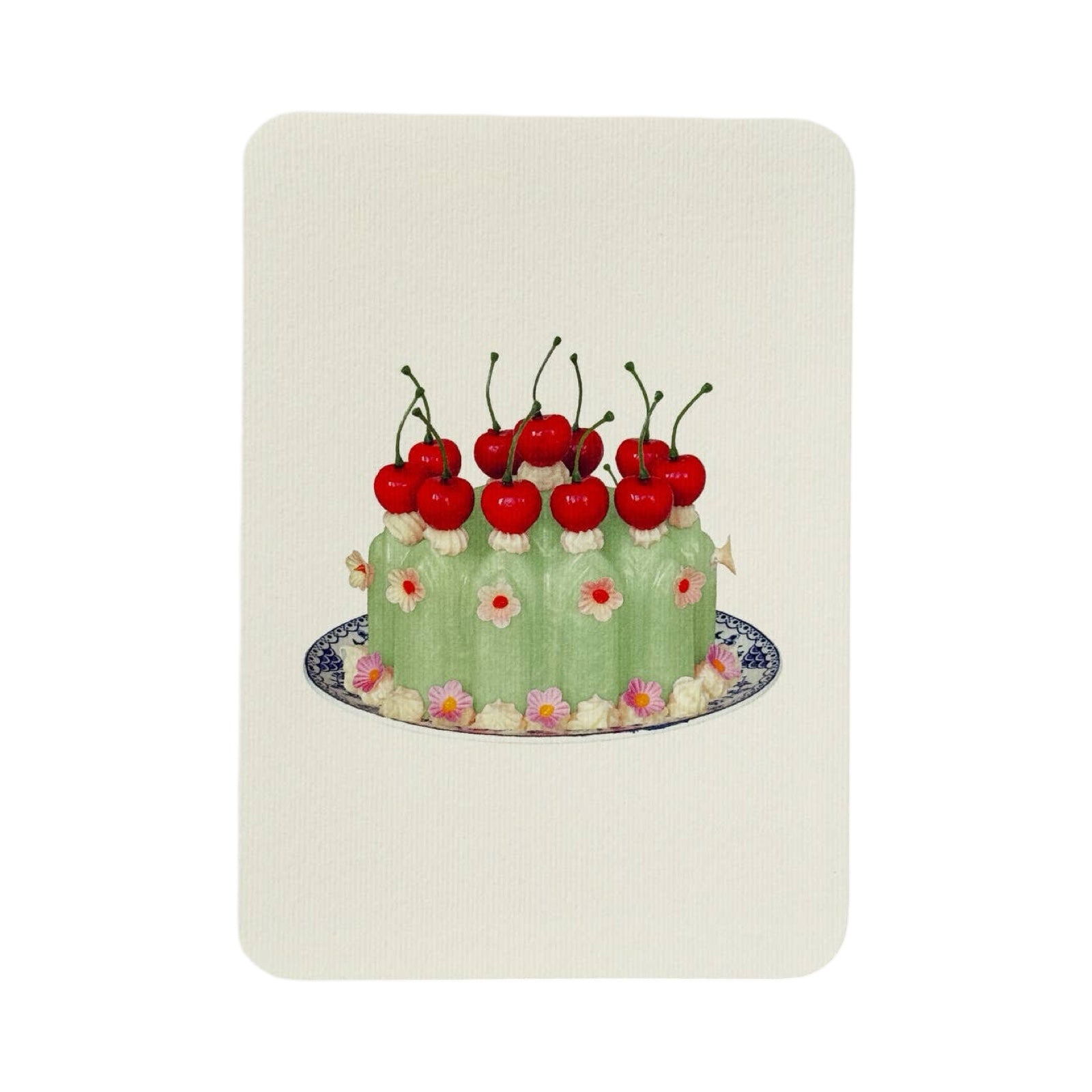 A cream coloured birthday card with a green coloured blancmange with cherries and cream on the top and small pink flowers, all on a blue and white plate.
