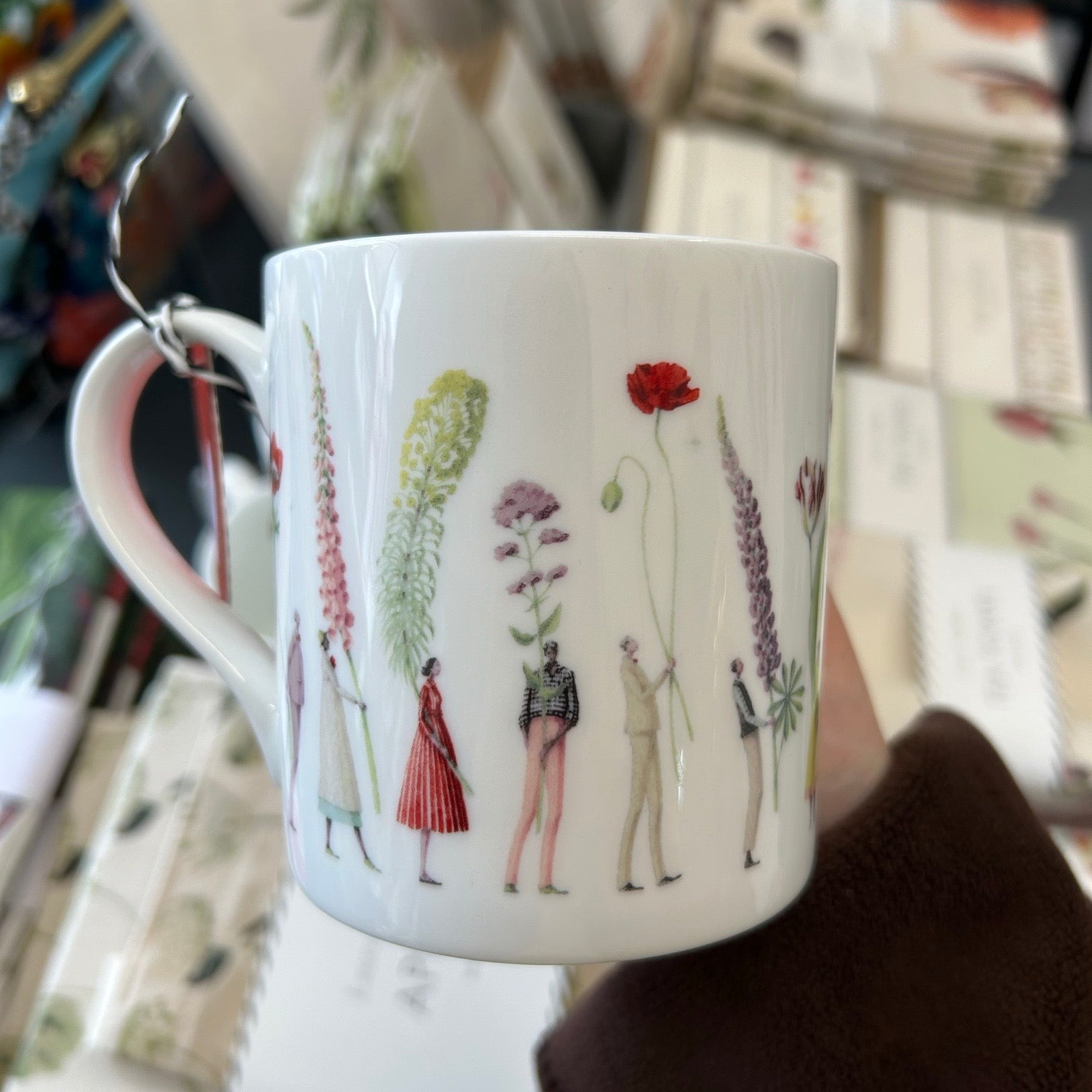 Mug with floral and figure design held by a hand in a blurred indoor setting.