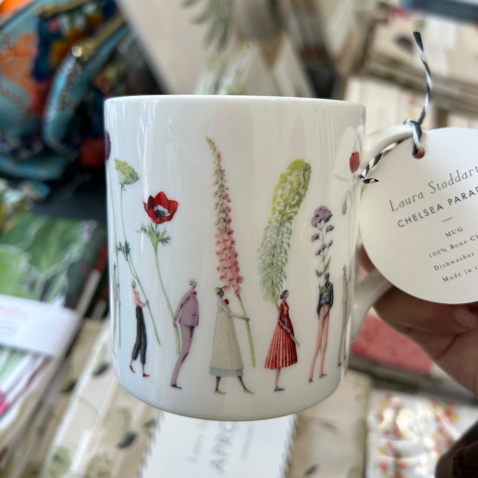 Mug with floral and figure design held by a hand, with a tag visible.
