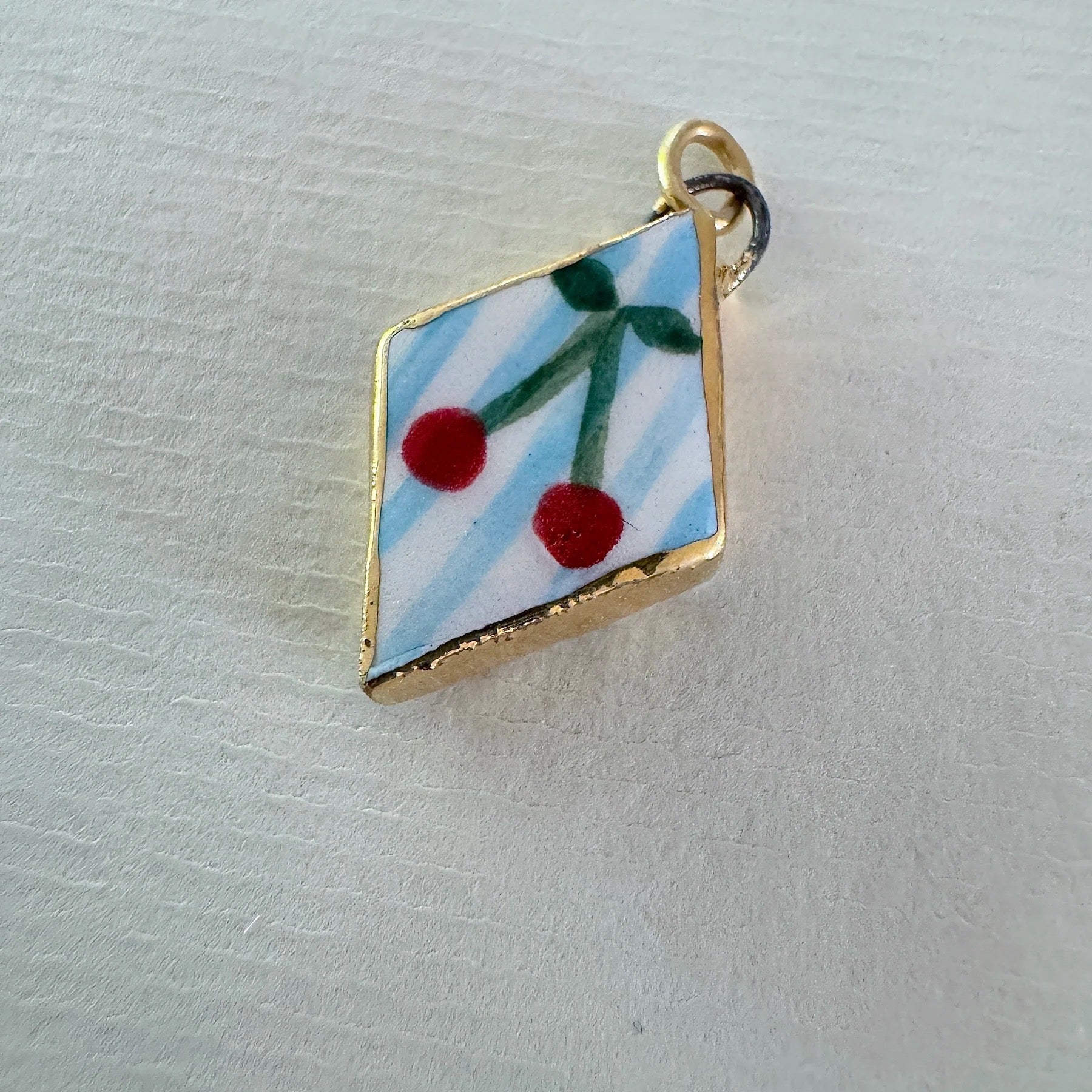 Diamond-shaped pendant with red cherries on a blue background, gold frame, on a light grey surface.