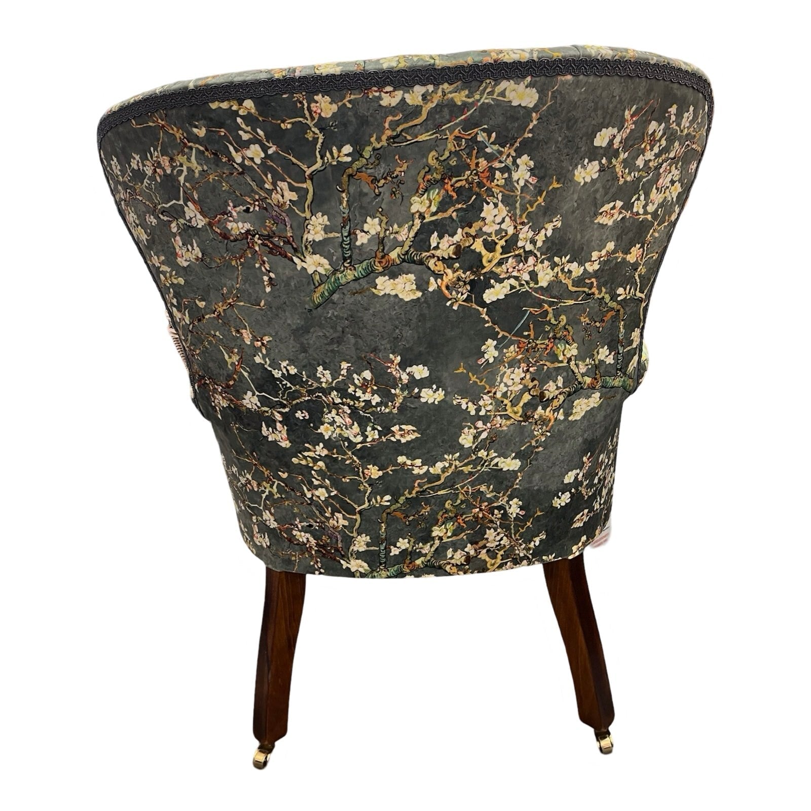 A grey velvet armchair with a floral pattern of white cherry blossoms, featuring button-back tufting, grey binding, and mahogany legs with gold castors.
