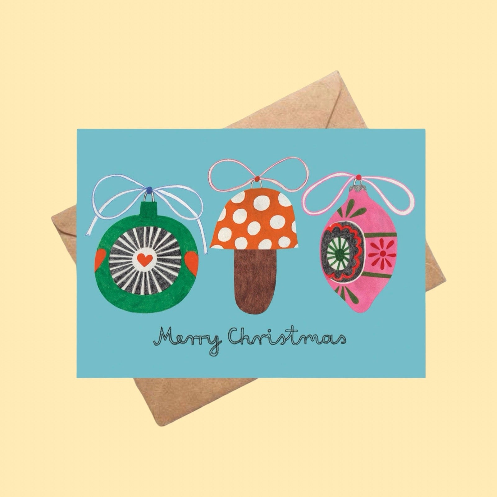 Christmas card with colorful ornaments and 'Merry Christmas' text on a light blue background, on a brown envelope on a yellow background.