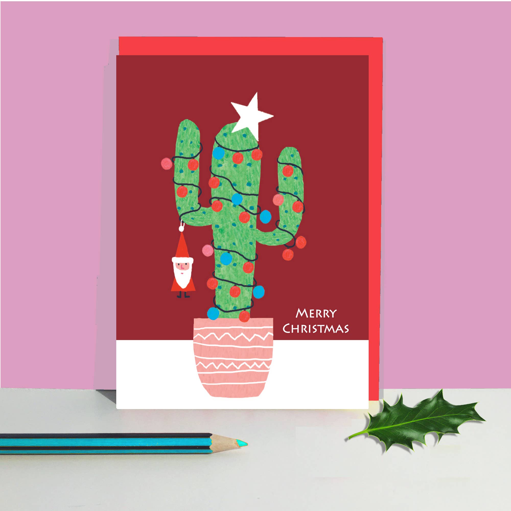 A Christmas greeting card featuring a green cactus decorated like a tree with a white star on top, colourful ornaments, a small Santa hanging ornament, and the words 'Merry Christmas' on the right. The card is shown standing upright on a white surface against a pink background, with a blue pencil and a green holly leaf nearby.