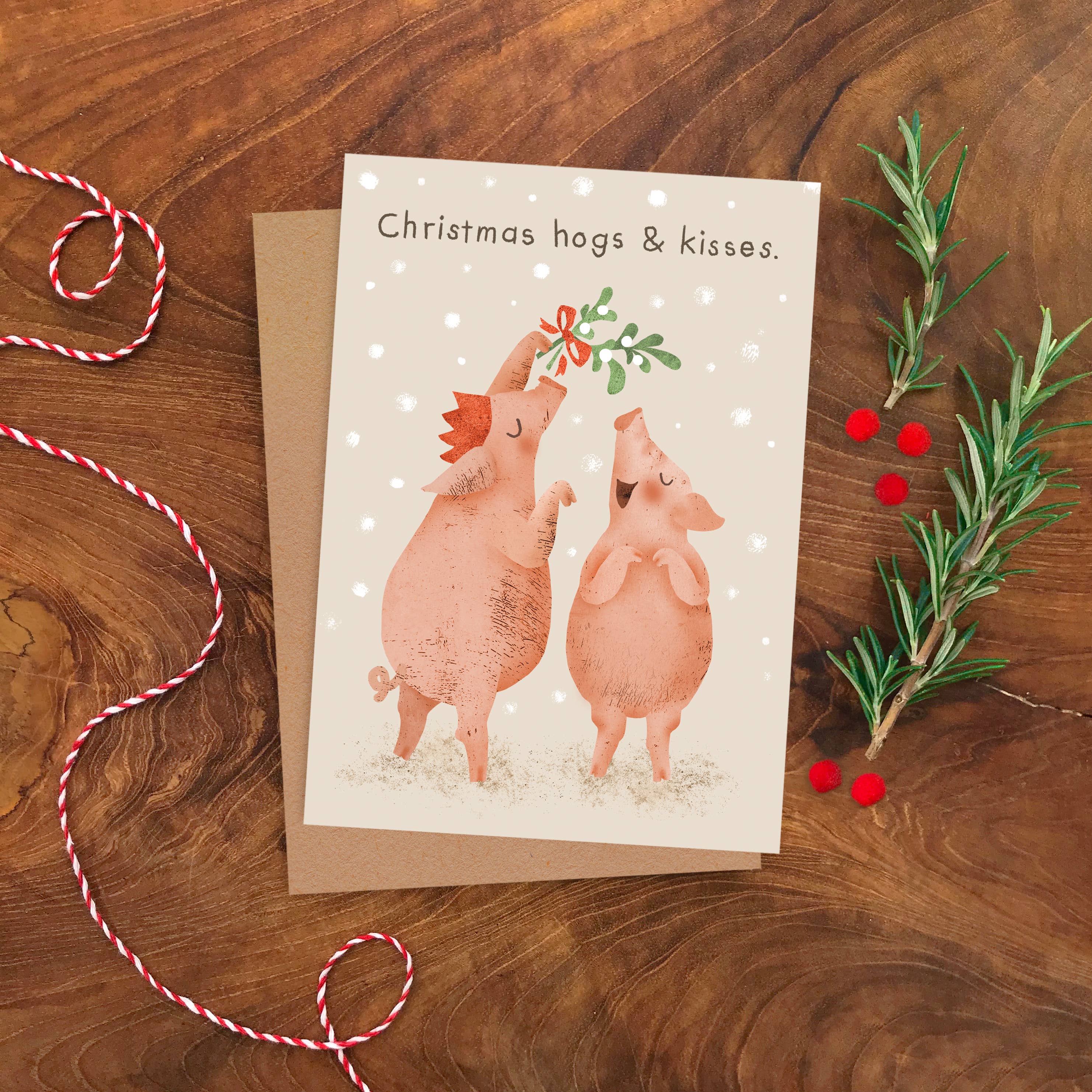 A Christmas greeting card featuring two cartoon pigs under a mistletoe branch, placed on a brown envelope on a wooden surface with red-and-white twine and greenery props.
