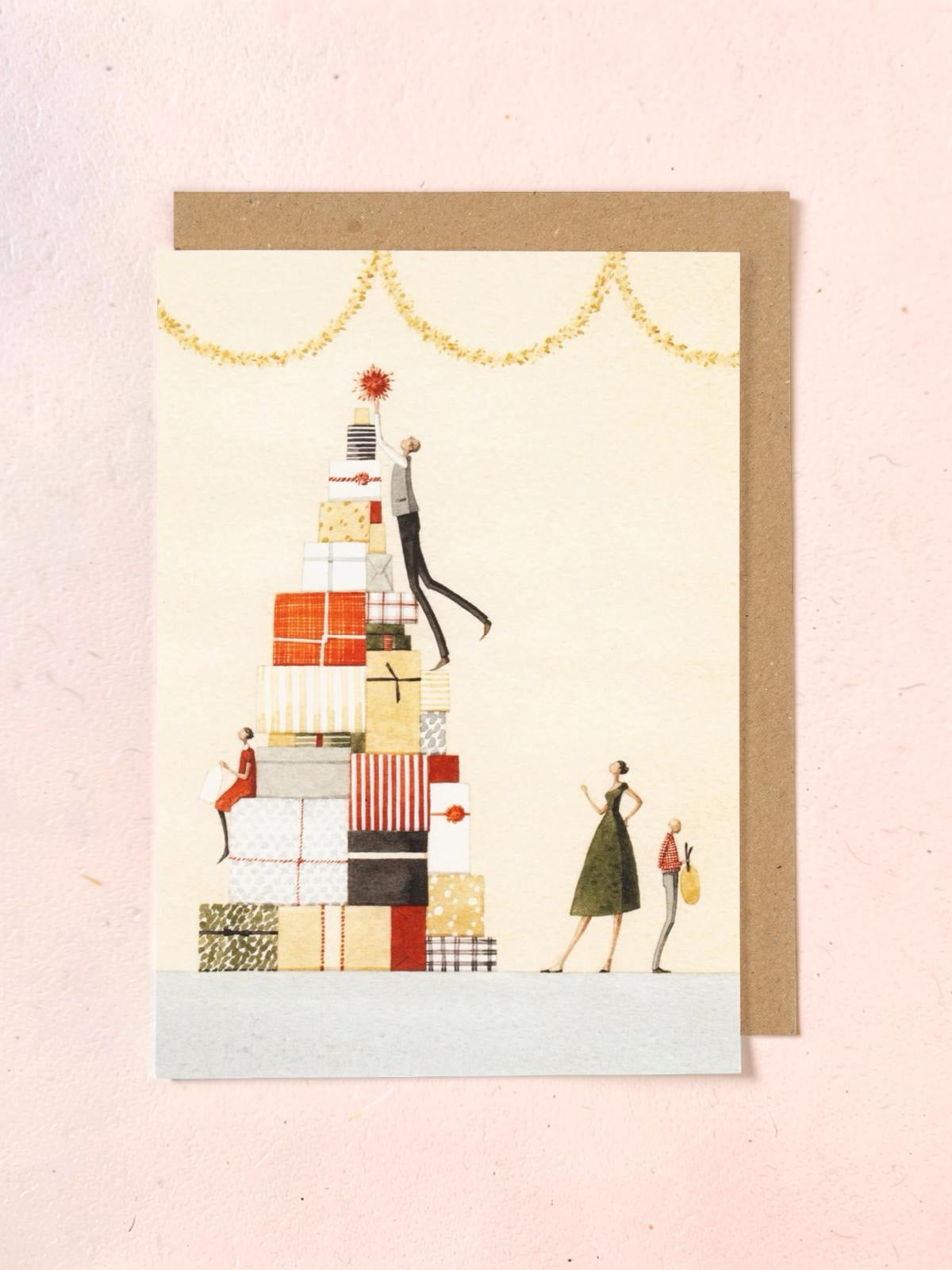Greeting card with illustration of people around a tall stack of presents on a light pink background.