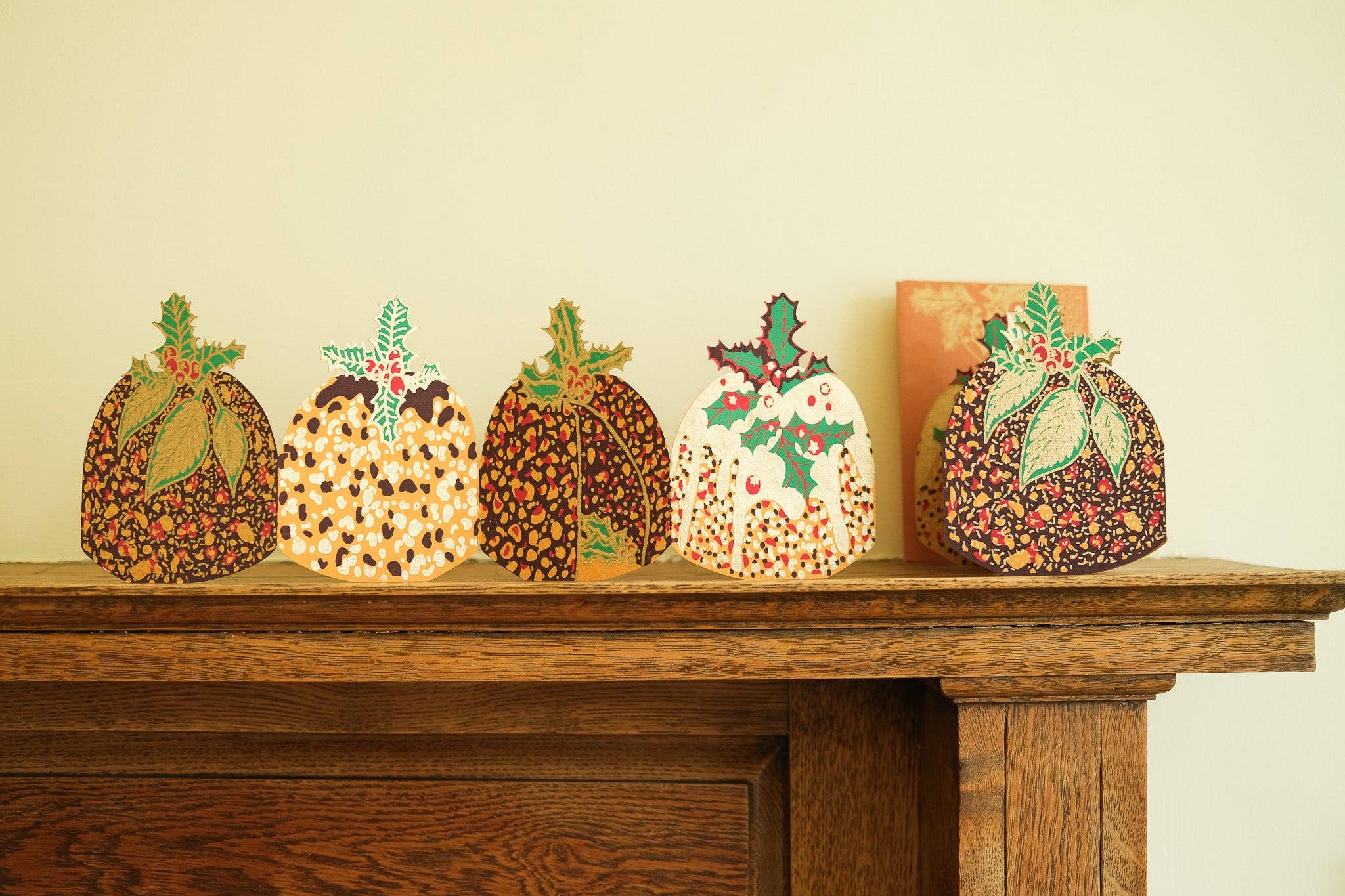 Five decorative Christmas puddings lined up on a wooden mantelpiece against a light beige wall.