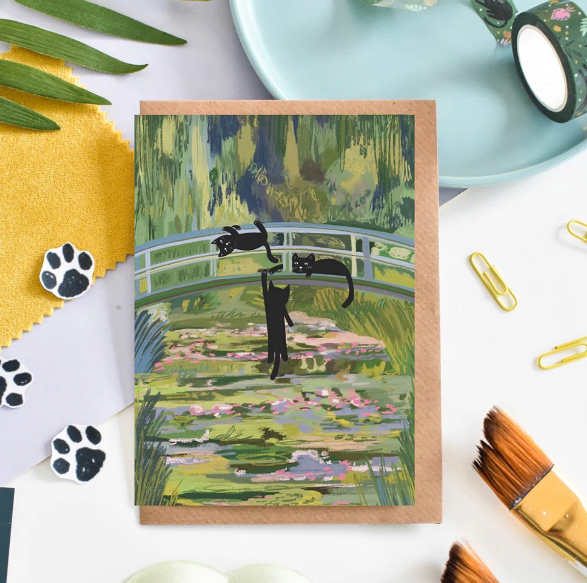 Greeting card with black cat illustration on a garden bridge, surrounded by stationery items.