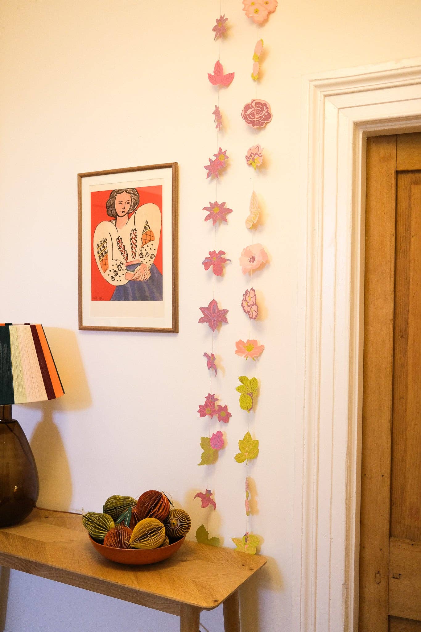 Decorative colorful paper flowers hanfing on a wall with a framed picture, and a wooden table with a bowl of fruit.