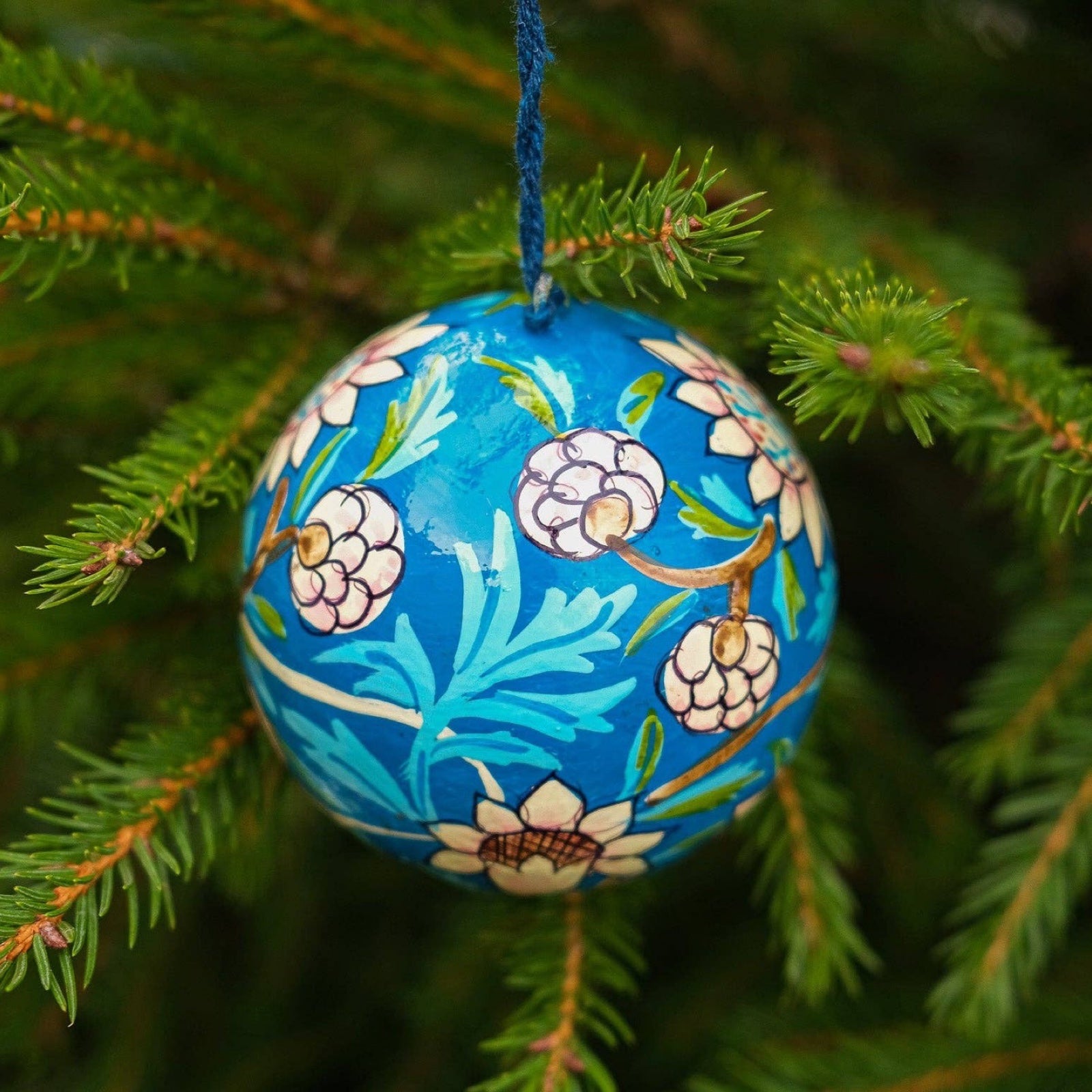A round papier mache bauble in cobalt blue with a white, turquoise and brown floral design hanging on a Christmas tree.