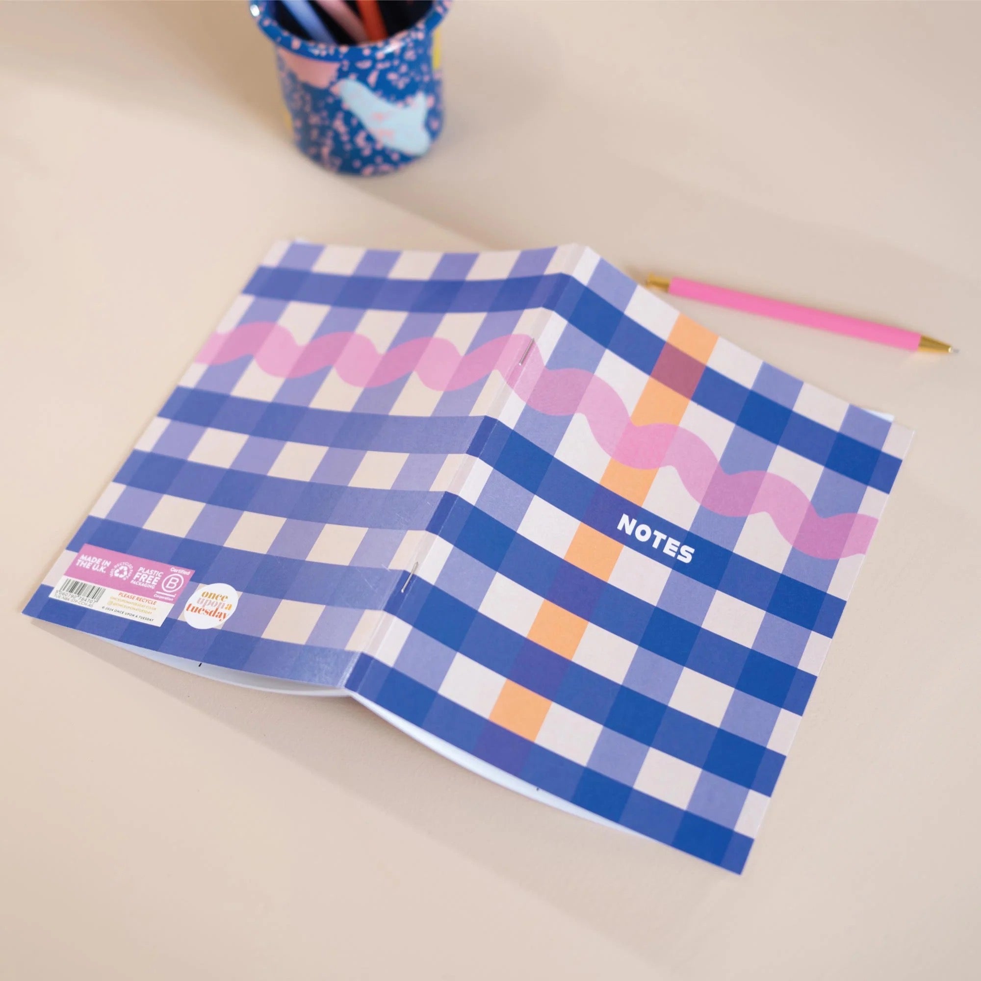 Open notebook with blue and pink checkered pattern on a beige surface, with a pen holder in the background.