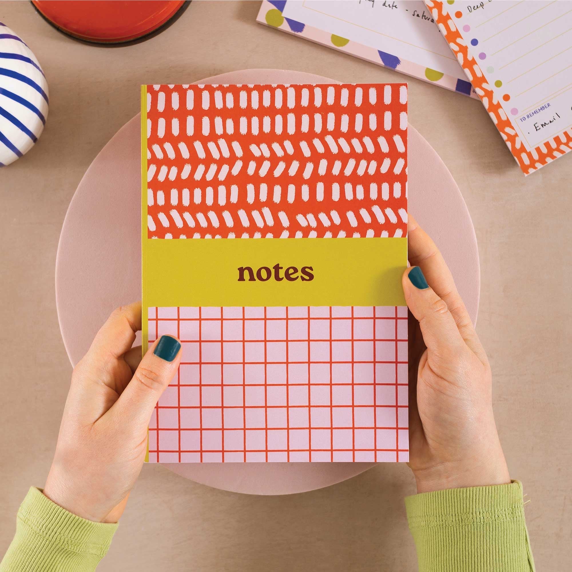 Person holding a colorful notebook with a grid pattern and 'notes' label on a beige surface.