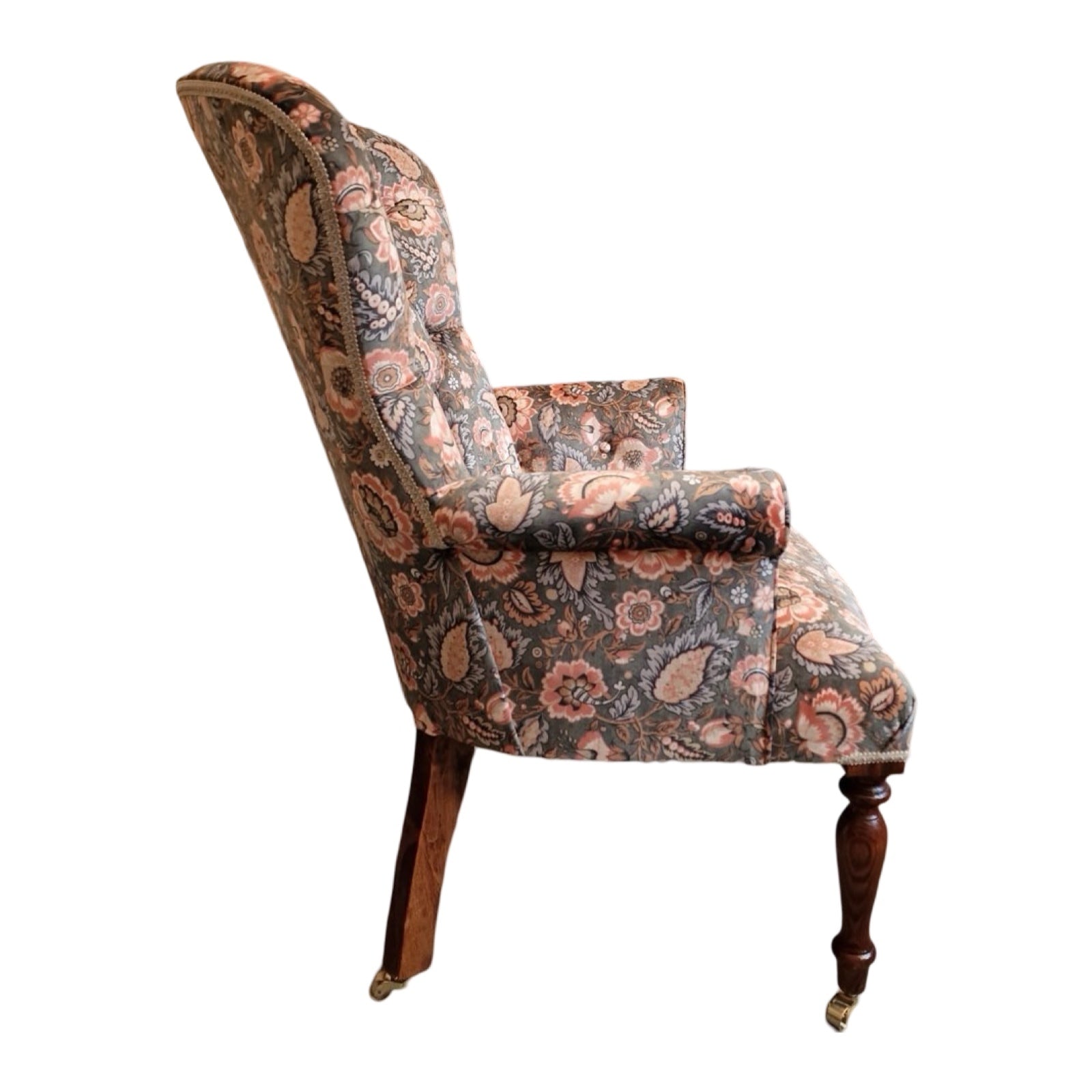 A luxurious armchair upholstered in light green/grey velvet with a floral pattern of coral flowers and pale blue leaves, featuring a button-back design, cream binding, and mahogany legs with gold castors.