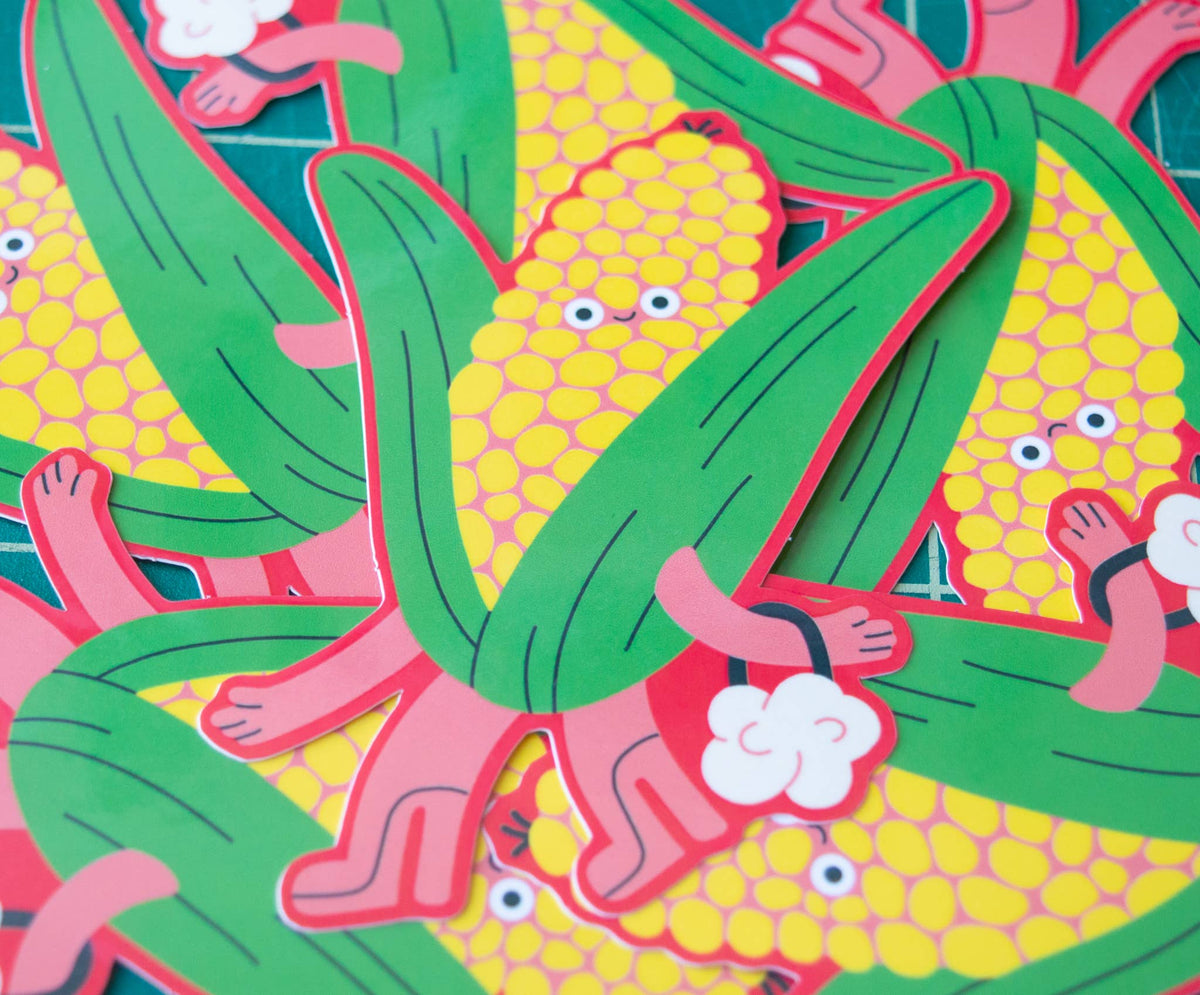 A close-up of colourful cartoon corn stickers or paper cutouts layered on top of each other. The design shows yellow corn kernels with eyes and smiling faces, green leaves, and pink stalks.