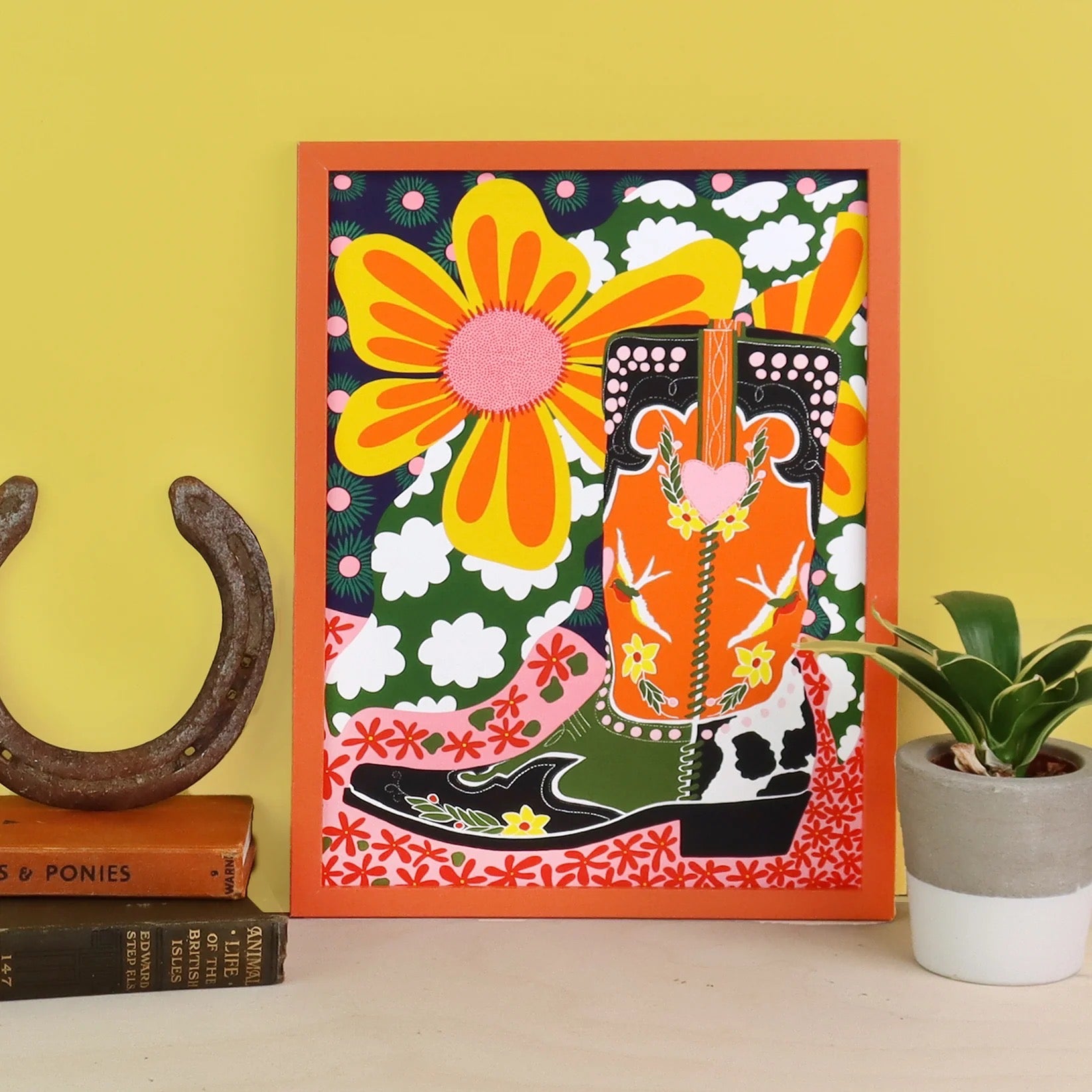 Framed colorful artwork with a cowboy boot and floral design on a yellow wall.