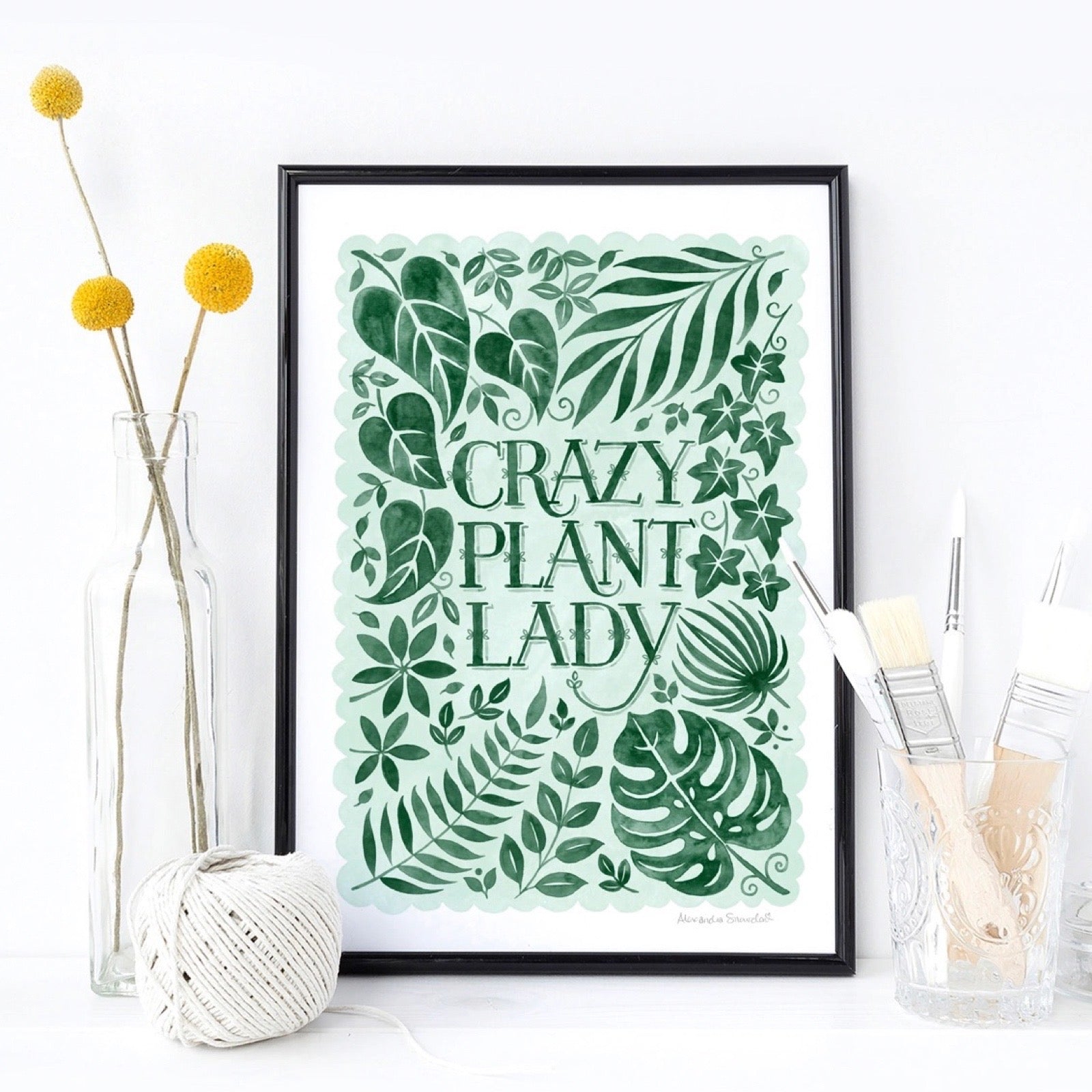 Framed artwork with 'Crazy Plant Lady' text and green leaf patterns on a light background, with plants and paint brushes.