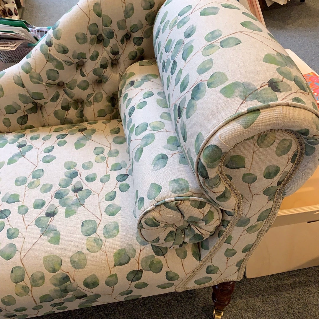 A handcrafted chaise lounge with cream leaves cotton fabric, mahogany stained legs, and brass-colored castors.