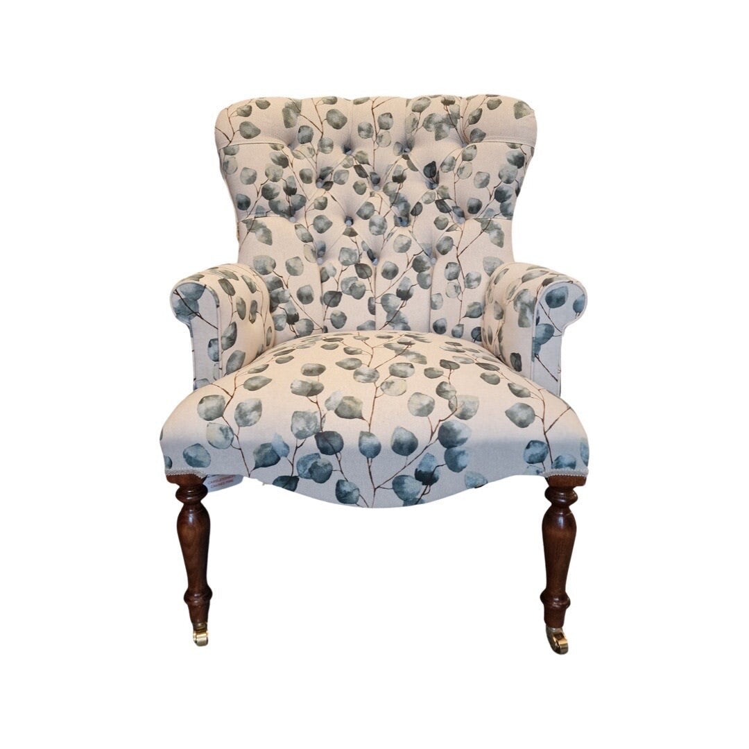 A hand-crafted cream linen armchair with green leaf pattern, button-back, cream binding, mahogany stained legs, and gold colored castors.