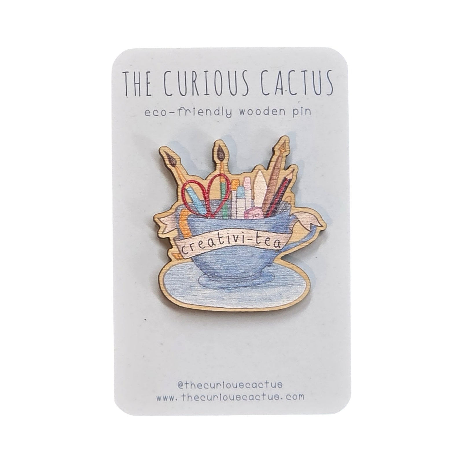 Wooden pin badge featuring a blue teacup filled with pens, pencils and scissors with the text 'creativi-tea' in a white banner on backing card.