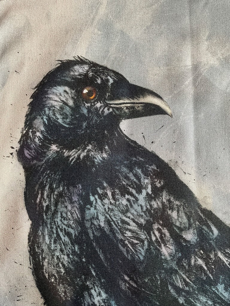 A tea towel featuring an illustration of a standing black crow, against a grey background.