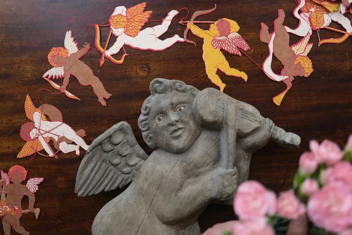 Stone statue of an angel with decorative cut-out cupids and flowers on a wooden surface.