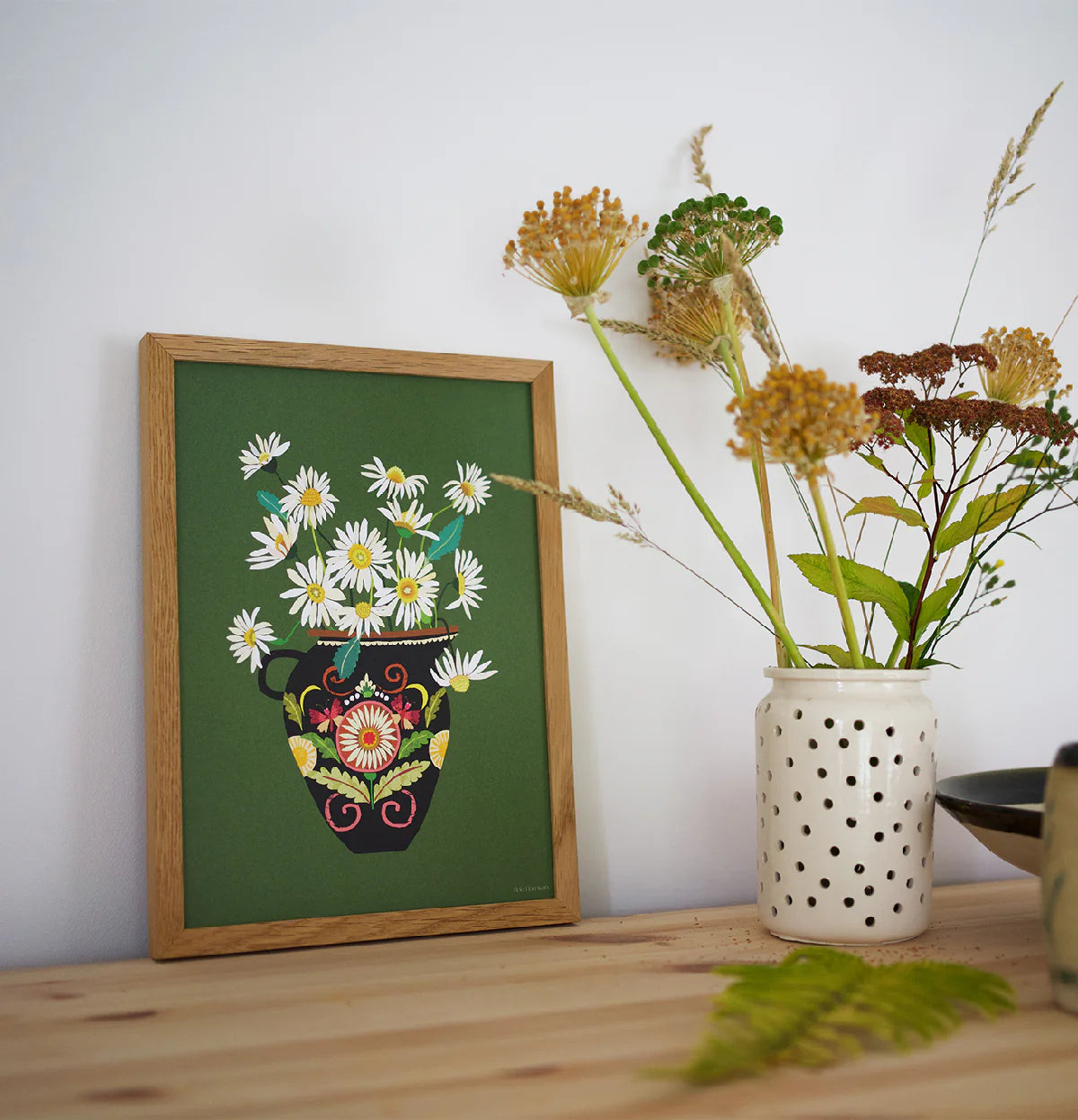 Framed artwork of flowers on a green background next to a vase with plants on a wooden surface.