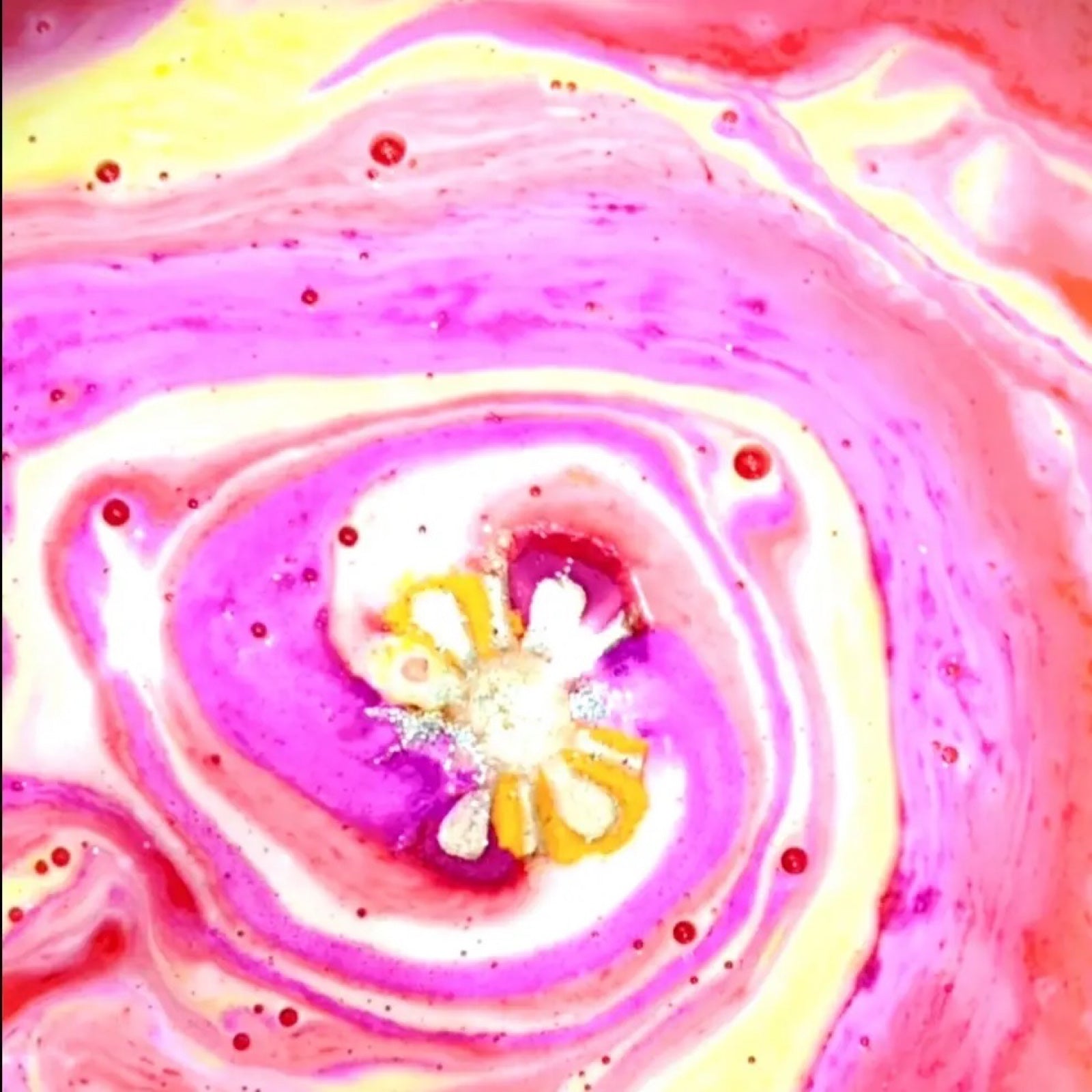 A pink and orange daisy-shaped bath bomb dissolving in the bath and turning the water pink, yellow and orange.