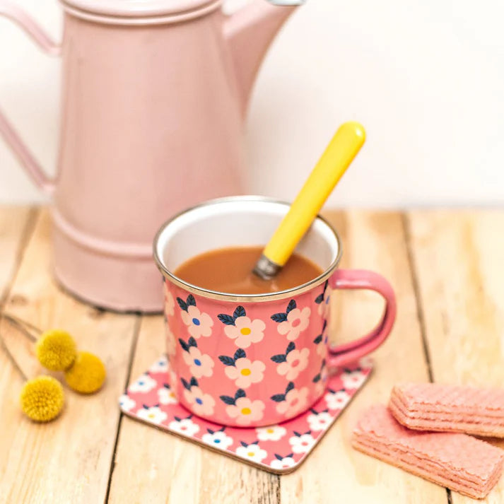 A pink enamel mug with a white and navy floral pattern sits on a matching coaster on a wooden table. A pink teapot is blurred in the background, with small yellow flowers and pink wafers.