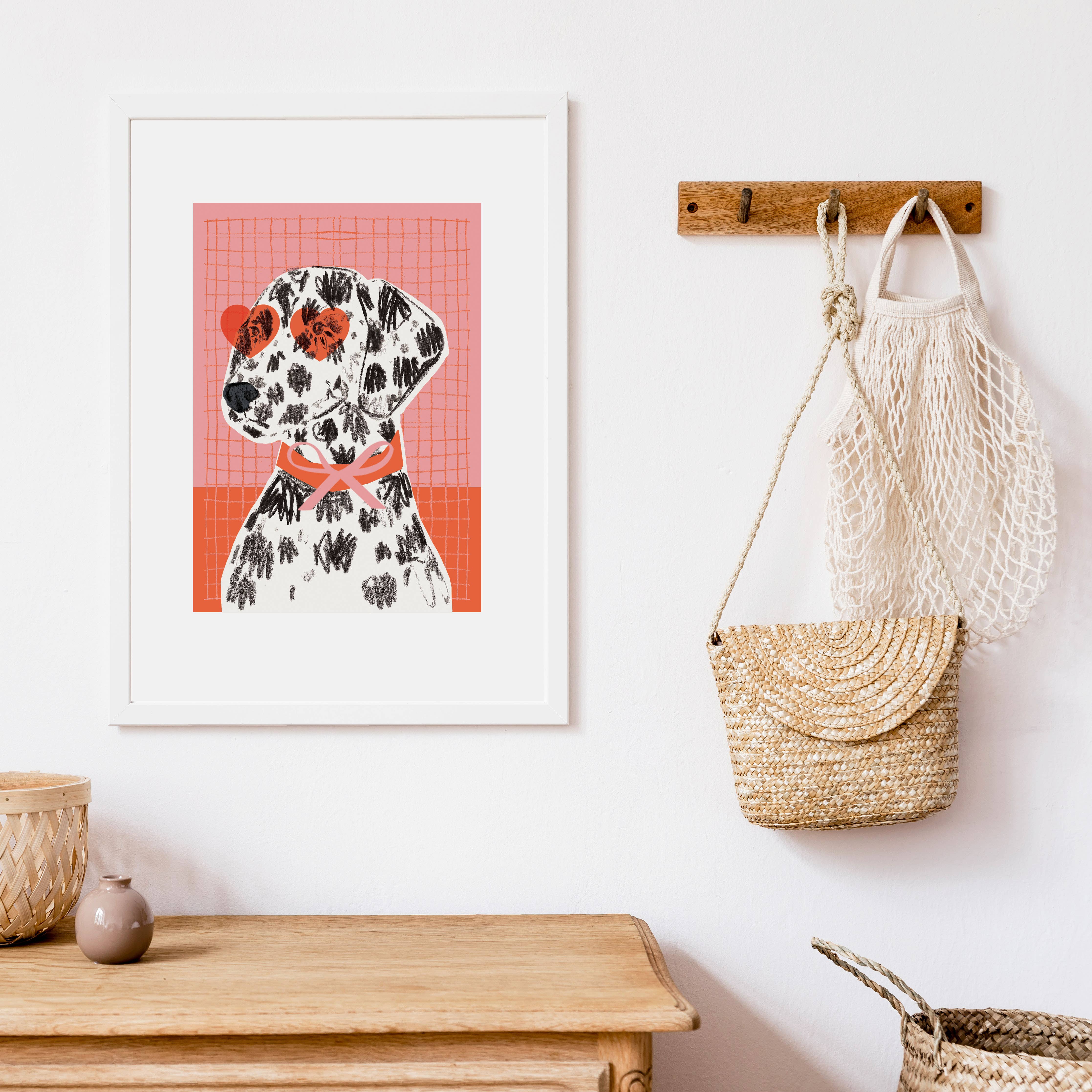 A framed art print featuring a stylised Dalmatian dog on a pink grid background, displayed on a white wall above a wooden bench. A wooden peg holds a cream mesh bag and a woven straw bag.