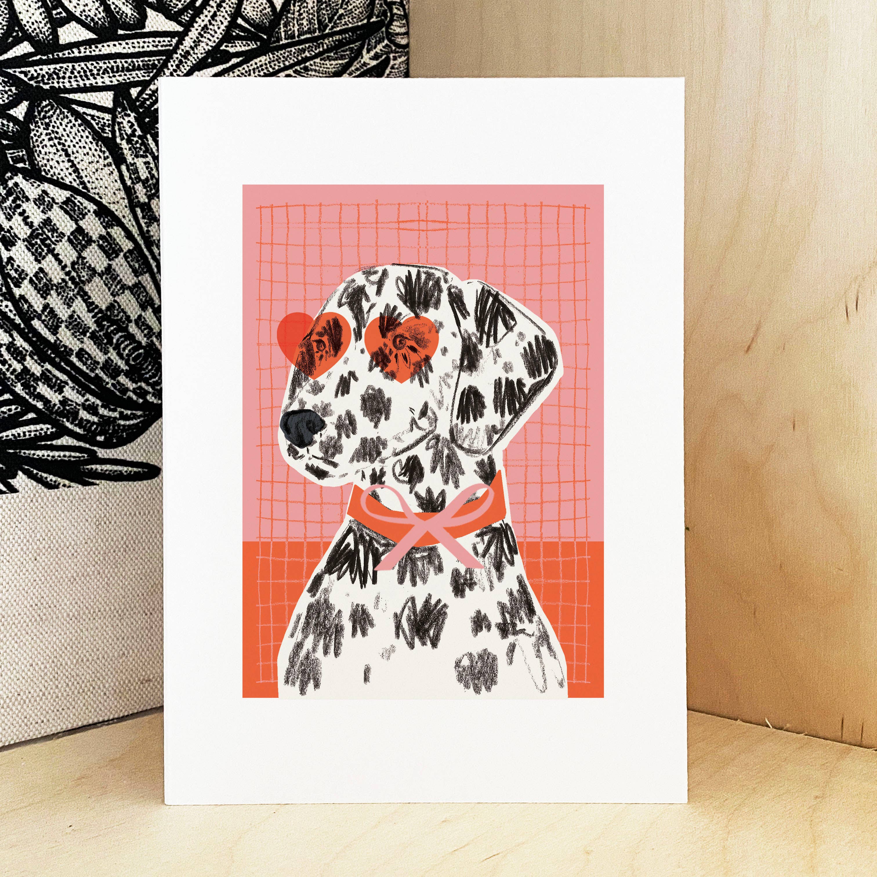 An art print card featuring a stylised Dalmatian dog illustration with black spots, red heart-shaped sunglasses, and a pink bow, displayed on a light wood surface.