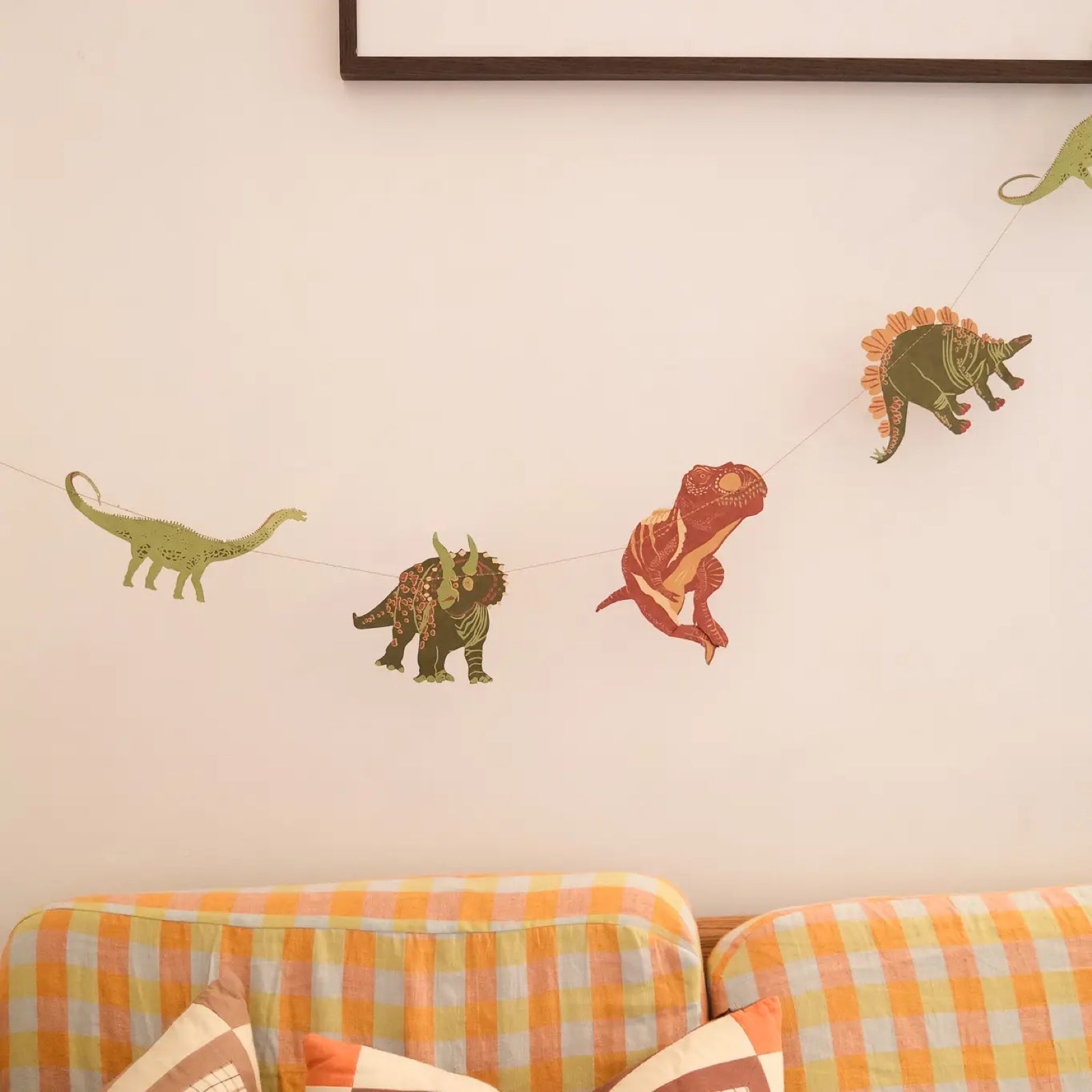 3m long dinosaur paper garland, featuring ten screen-printed dinosaurs including the tyrannosaurus, triceratops and stegosaurus hanging against a cream wall with a yellow checked sofa below.