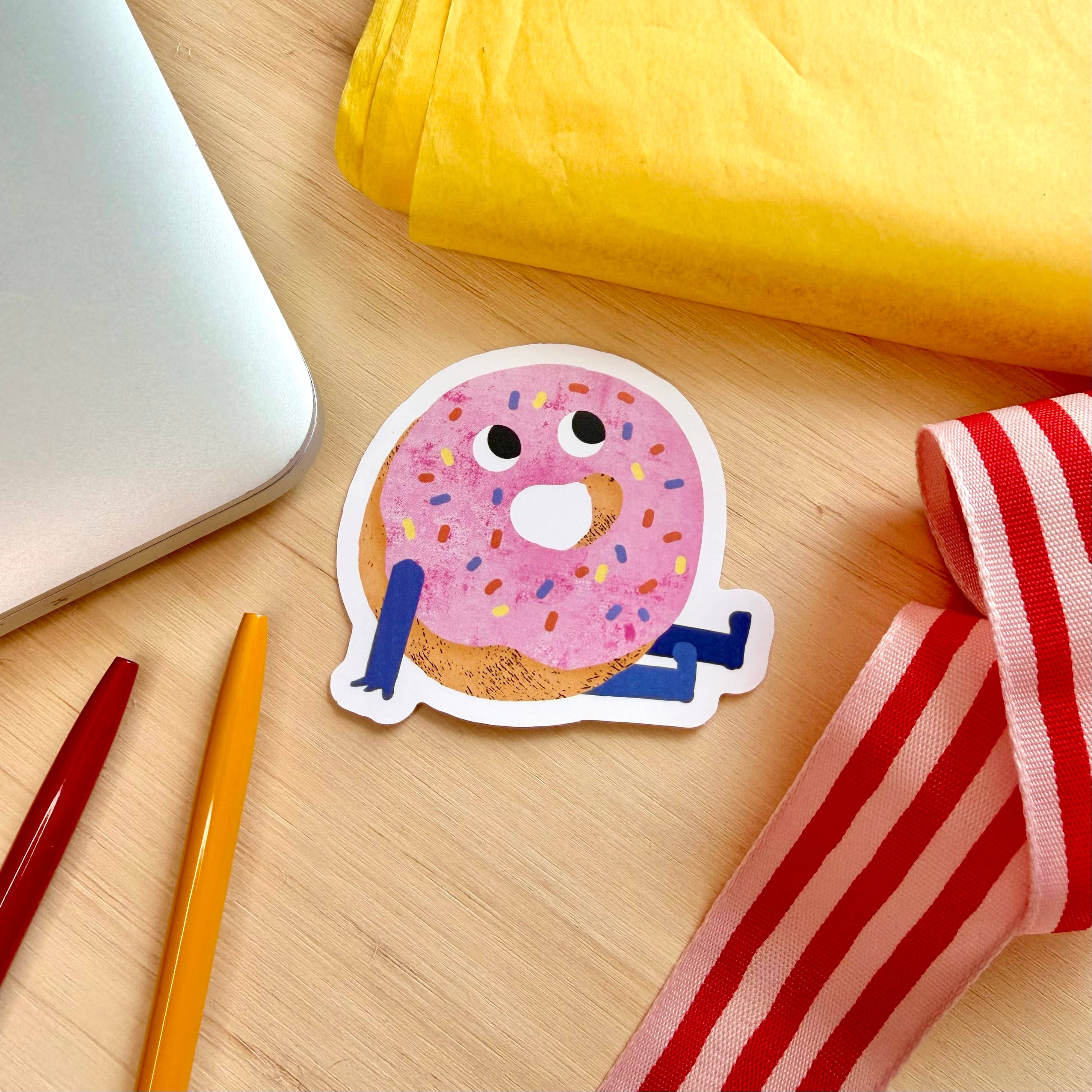 A die-cut sticker of a cute cartoon donut with pink frosting, colorful sprinkles, and blue limbs, placed on a light wood desk with a laptop corner, yellow paper, and red-and-white striped ribbon.