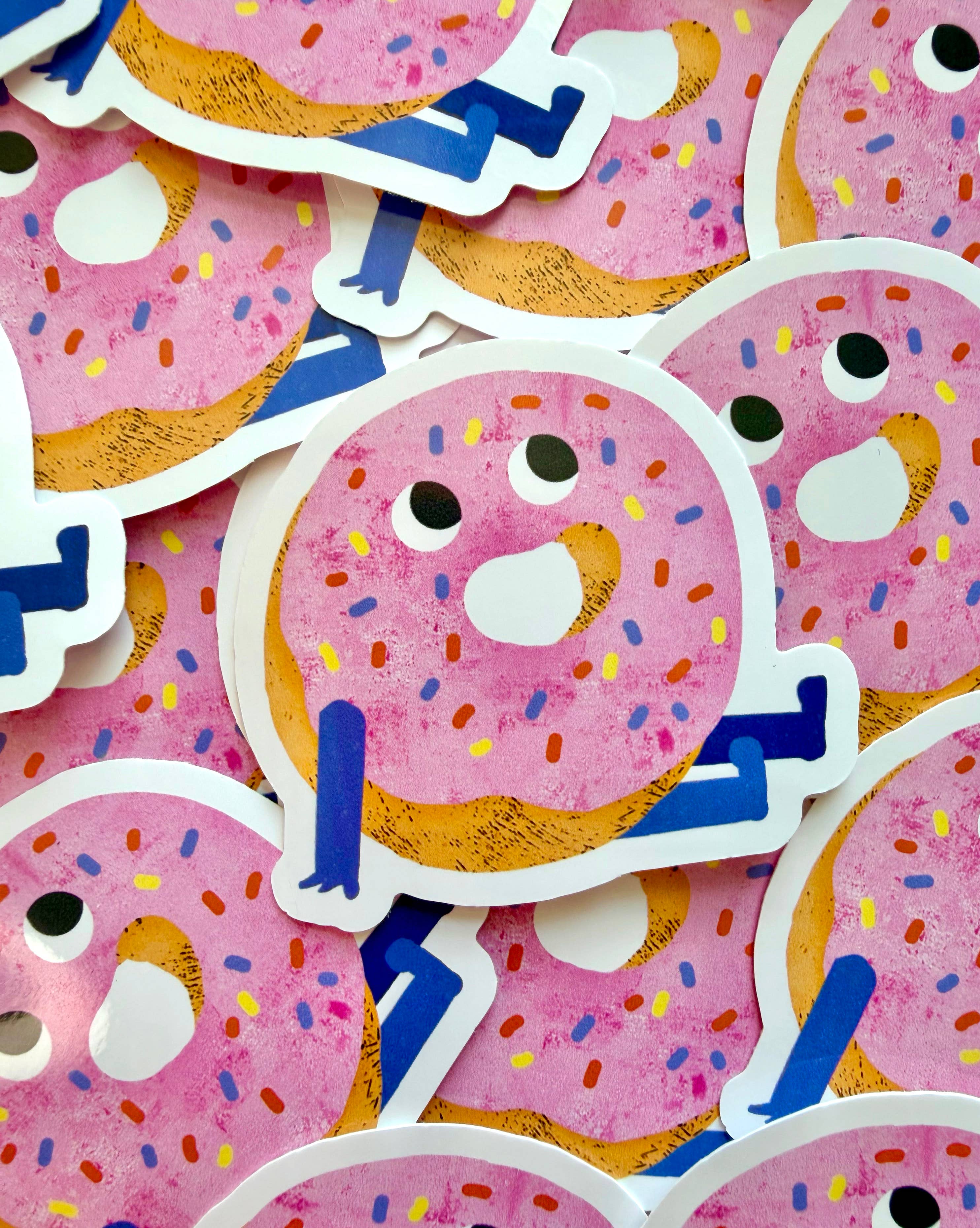 A pile of die-cut stickers featuring cute cartoon donuts with pink frosting, colourful sprinkles, and blue limbs on a pink background.