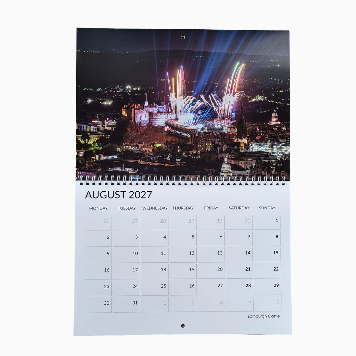 A spiral-bound desk calendar page showing August 2027, with a large nighttime city fireworks photograph at the top and a printed calendar grid below on a white page.