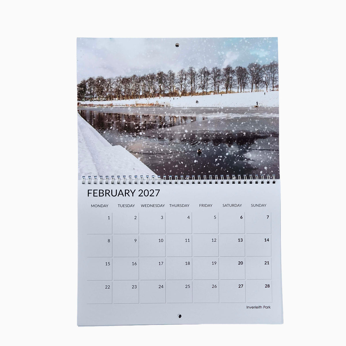 A spiral-bound February 2027 desk calendar page featuring a winter landscape photograph of a snowy river and trees, with a printed calendar grid below.
